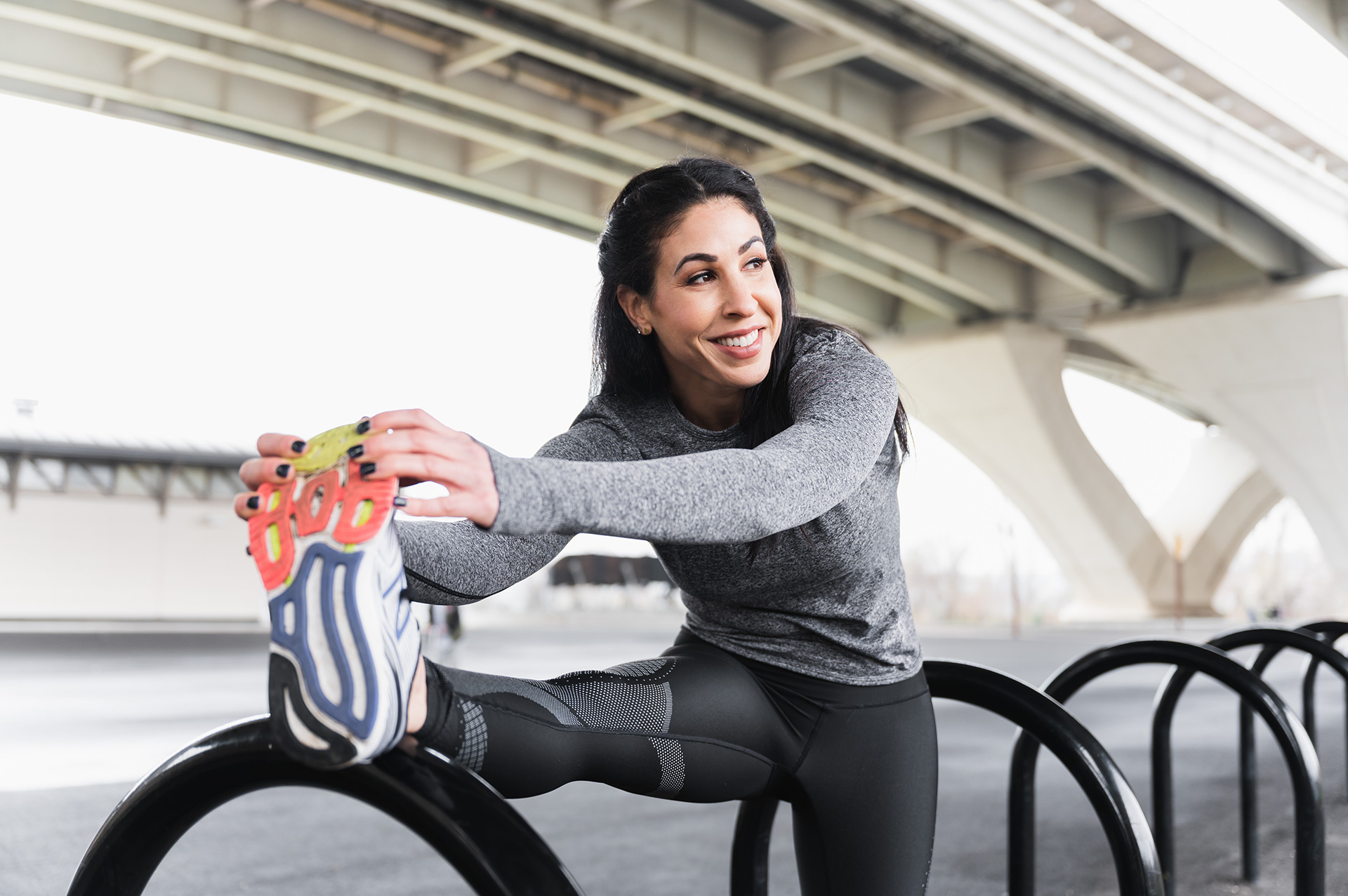 Woman stretching outdoors under a bridge, smiling, wearing athletic clothing, with a bike rack nearby.
