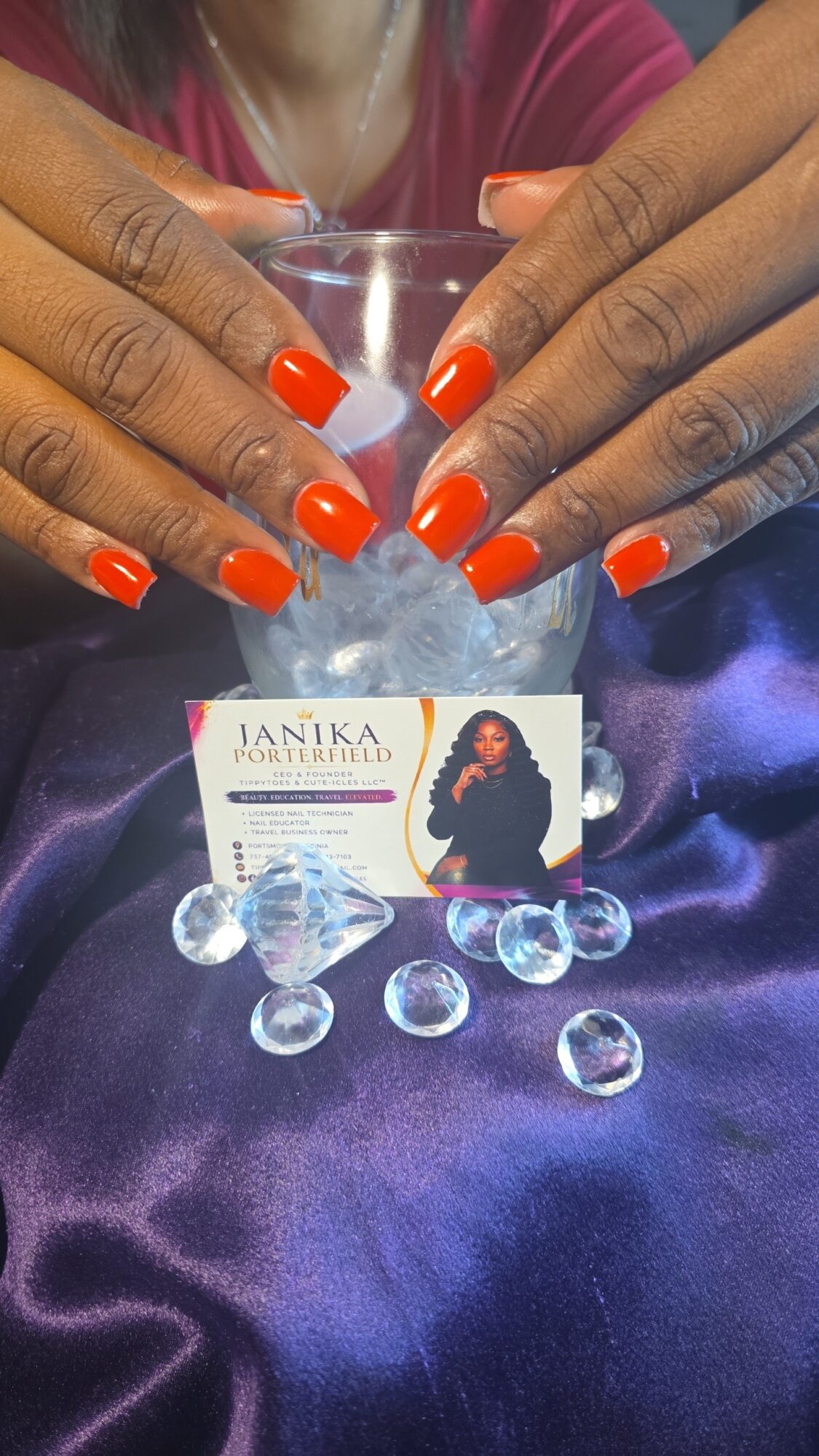 Hands with orange nail polish hold a glass with ice, on purple satin fabric, with a business card and ice cubes nearby.