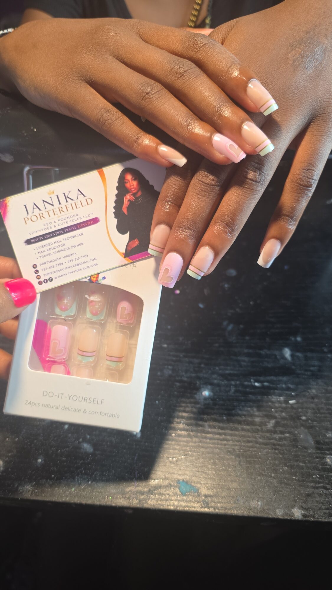 Two hands with manicured nails resting on a black surface, with a box of nail polish samples nearby.