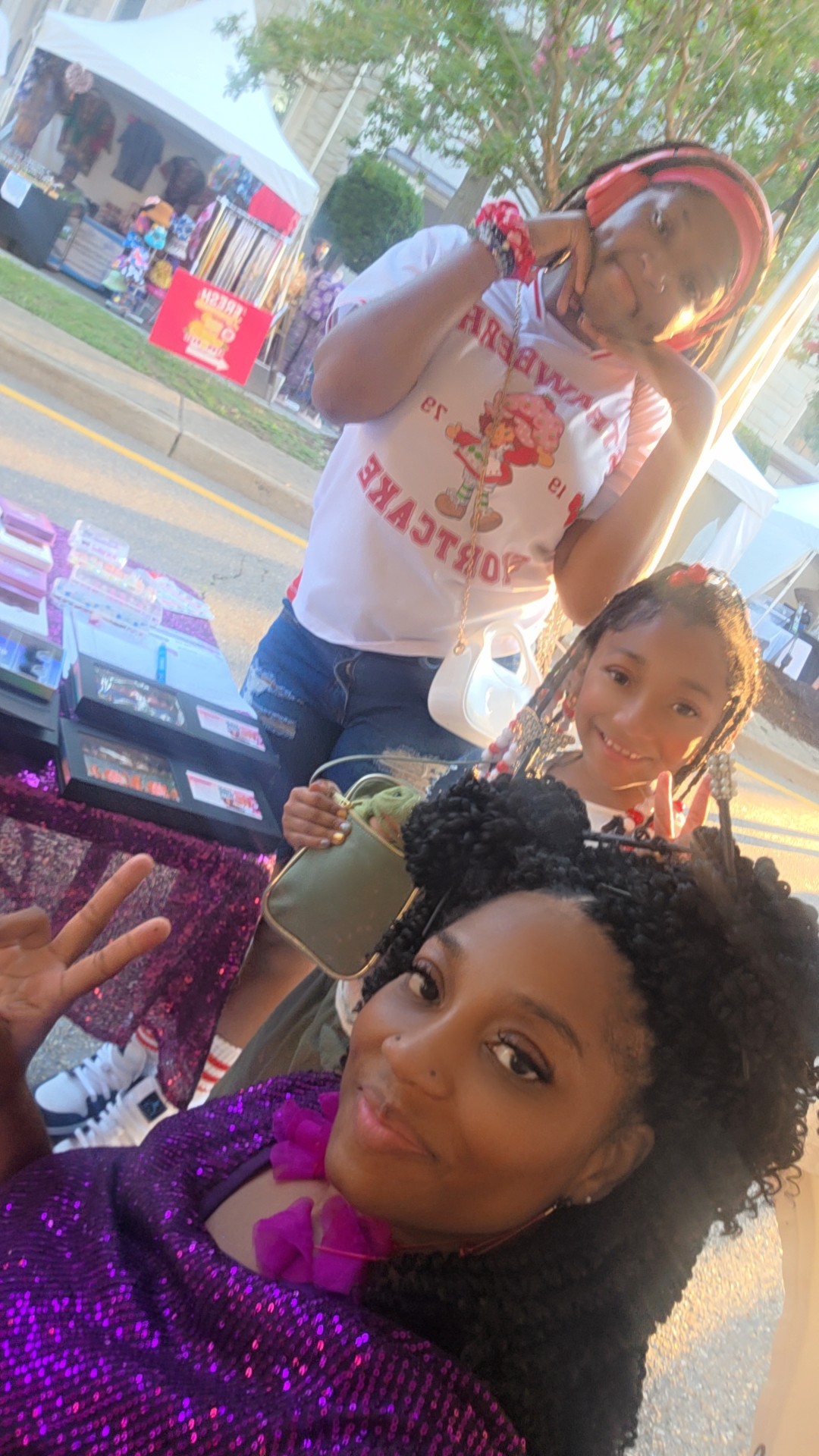 Three women posing outdoors near a table with books, with trees and a street in the background.