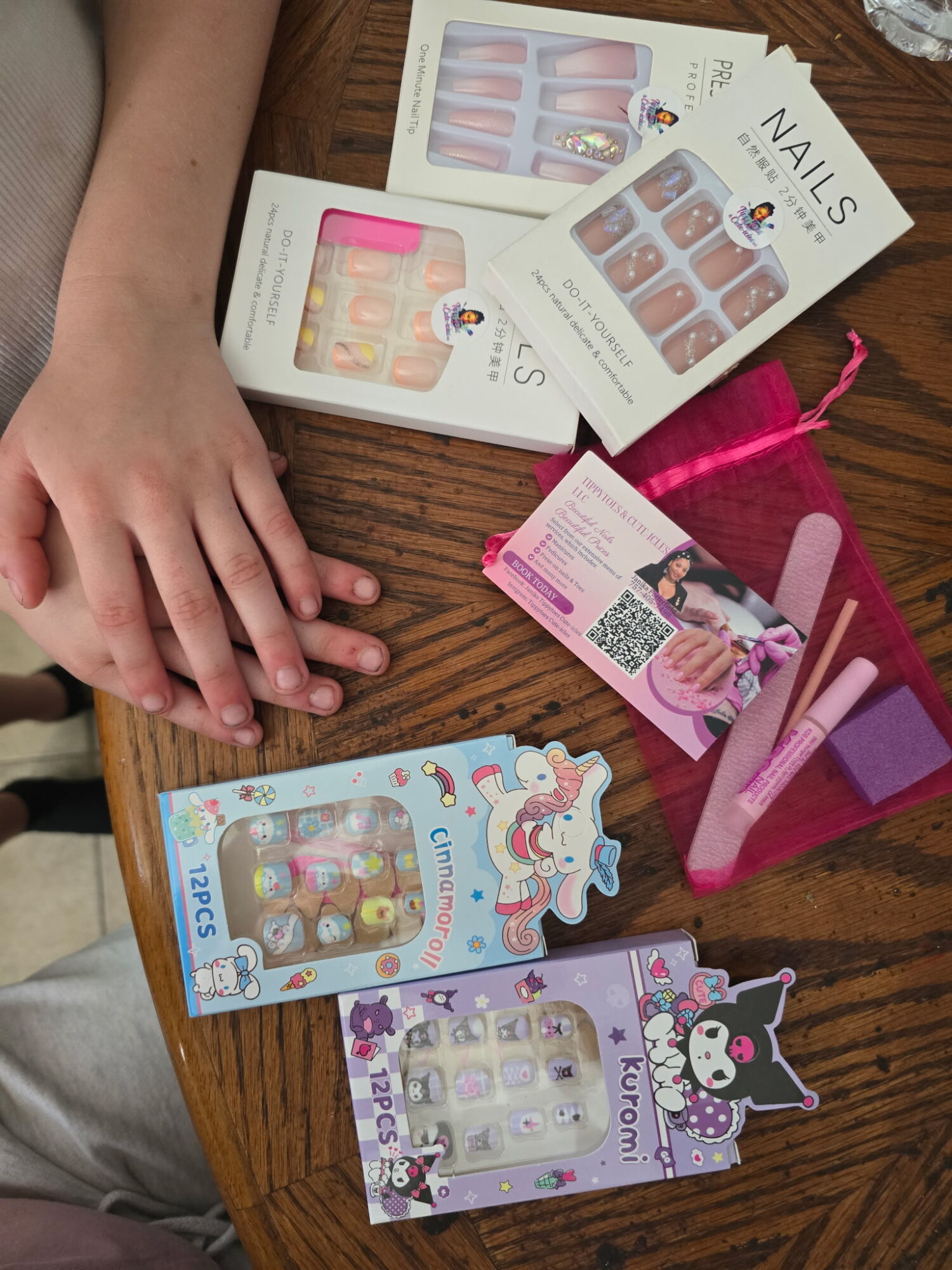 Hands resting on a wooden table with nail polish sets, pink tools, and a card, all related to nail art.
