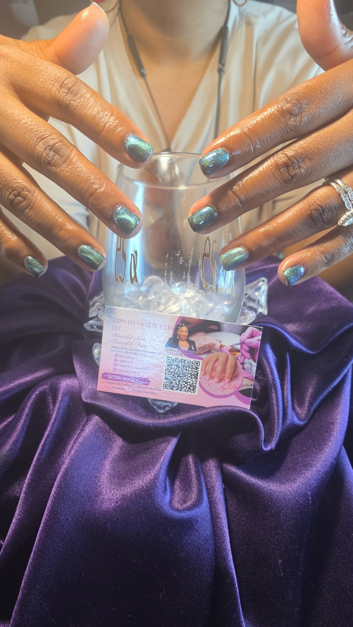 Person with multiple rings and painted nails holds a clear glass with a card underneath, resting on purple satin fabric.