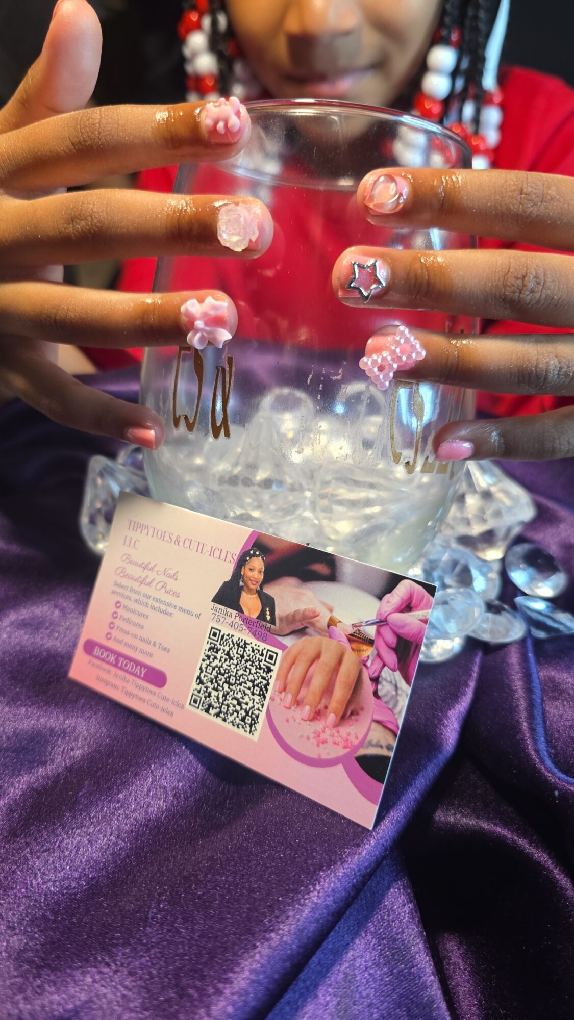 Person with decorated nails holding a glass bowl with ice, a card with a QR code, and purple fabric background.