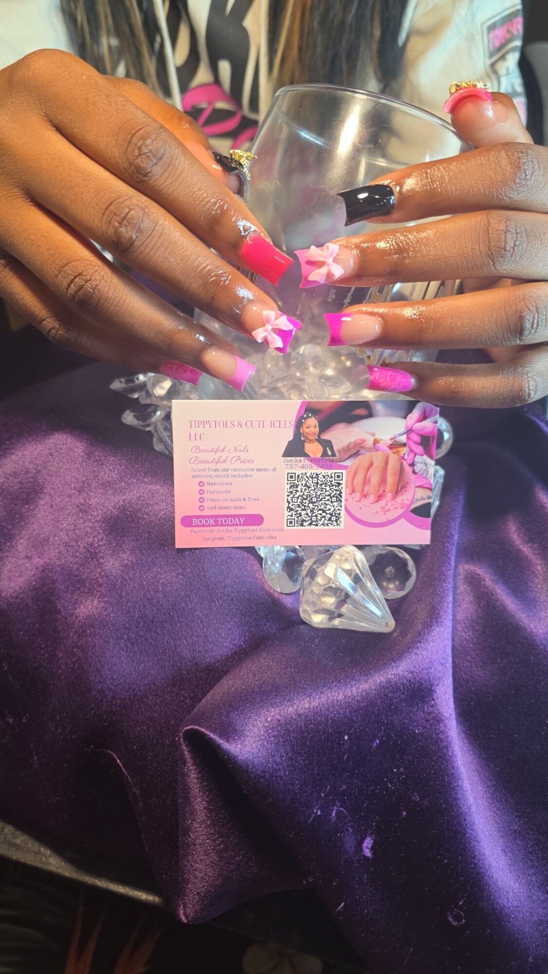 Hands with pink and white nail polish holding a glass, with a business card and purple satin fabric nearby.