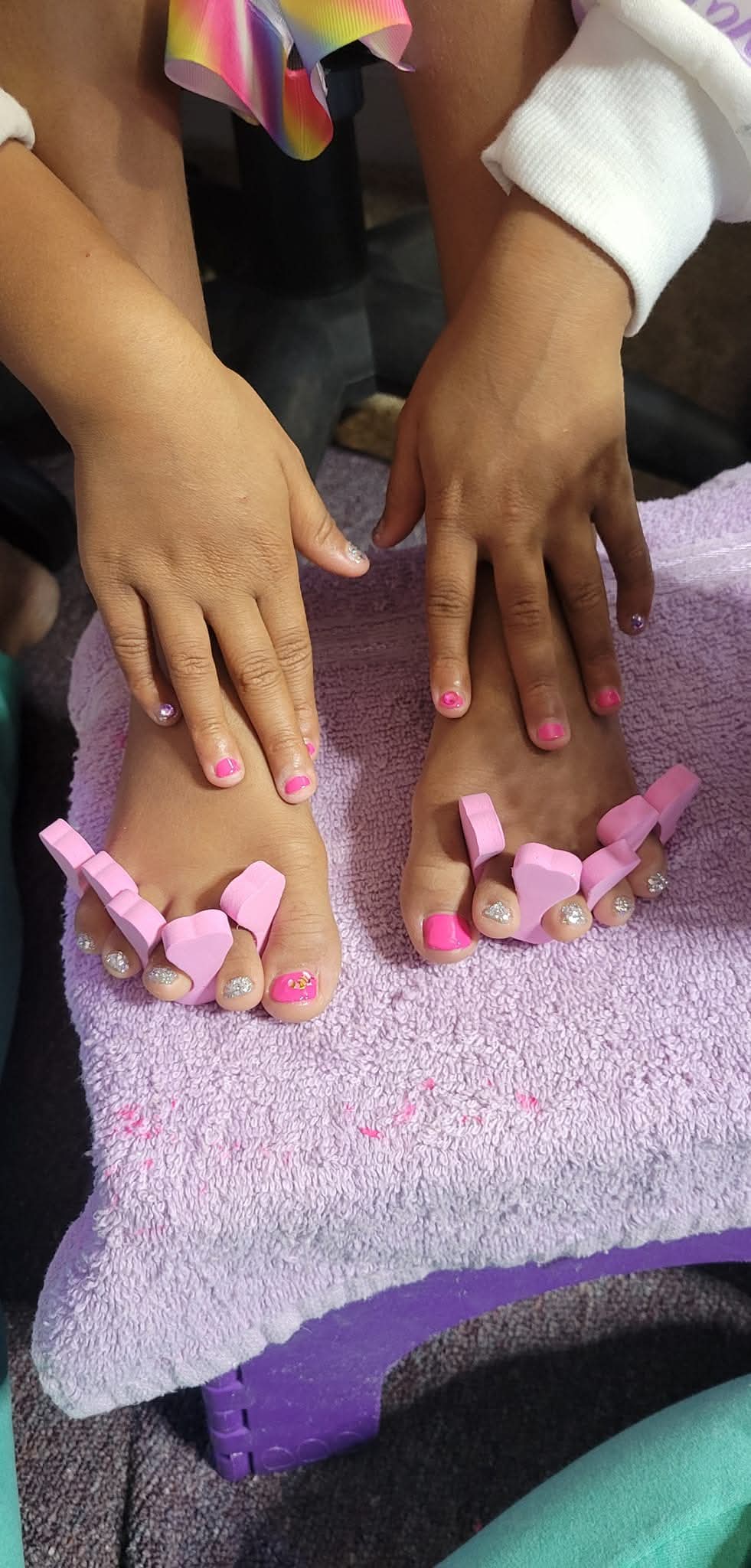 Two children have pedicures with pink nail polish, feet resting on a purple towel, hands visible, one child wearing a white sleeve.