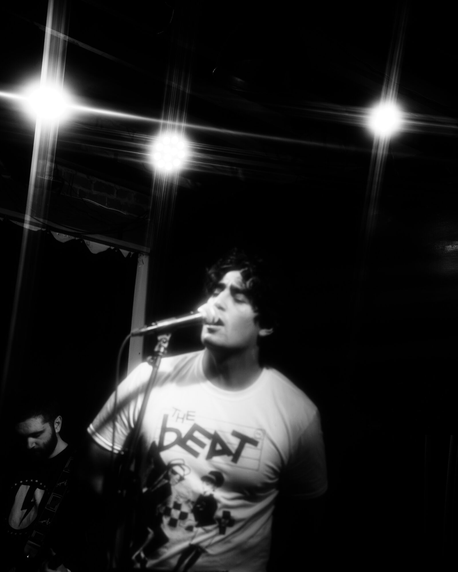 Person singing into microphone on stage with bright lights overhead, black background, and another musician partially visible.