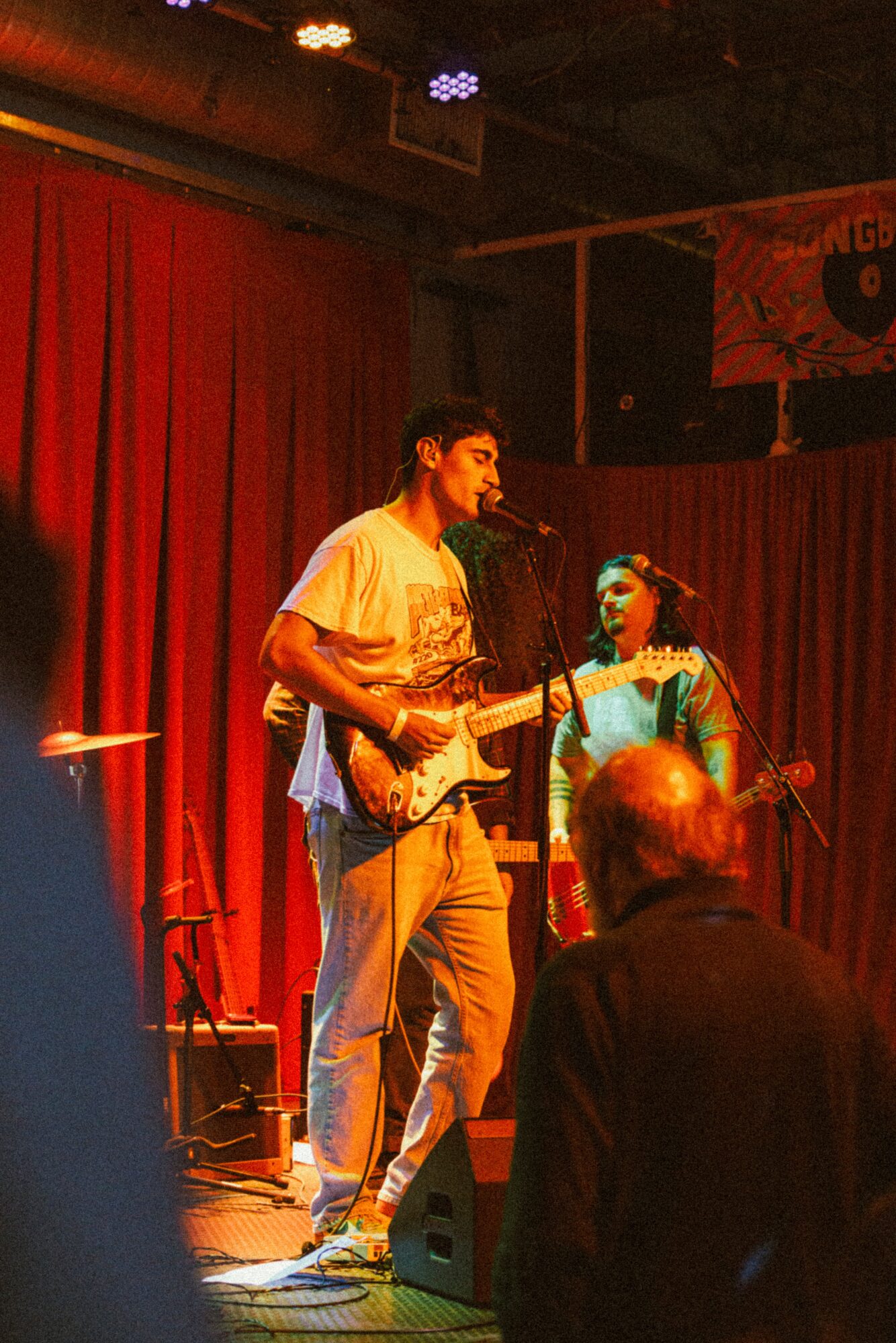Two musicians perform on stage with red curtains, one playing guitar and the other singing, audience members in foreground.