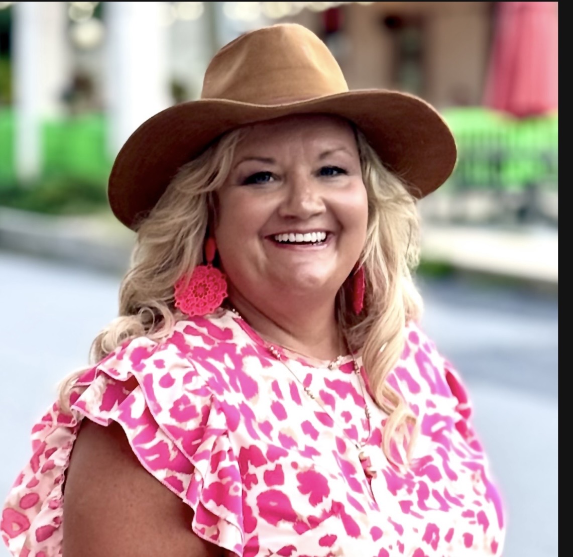 Smiling woman wearing a wide-brimmed hat and pink patterned dress outdoors.