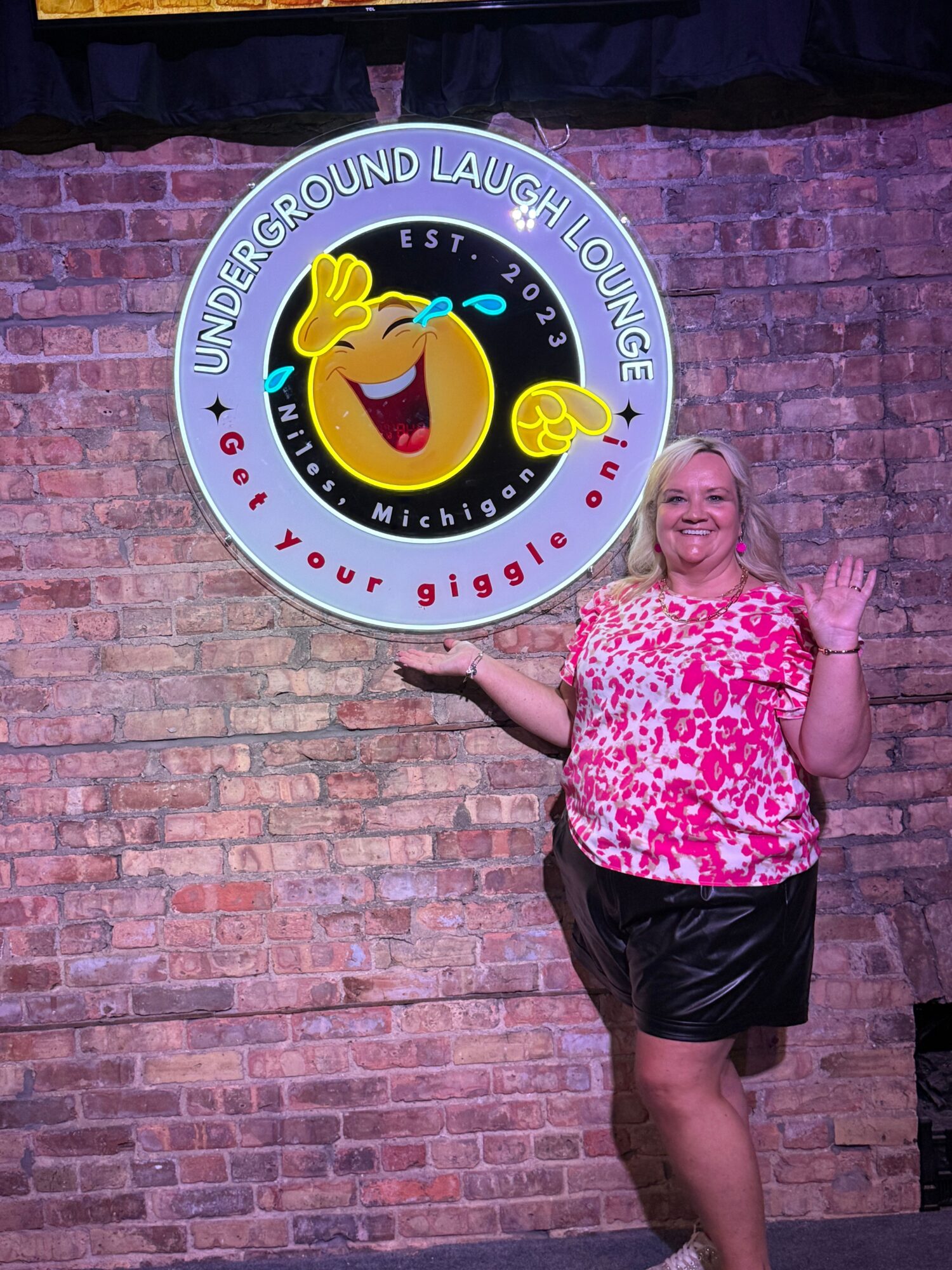Person lying on brick wall floor, smiling, wearing pink and white top and black shorts, next to a circular sign with a laughing emoji.
