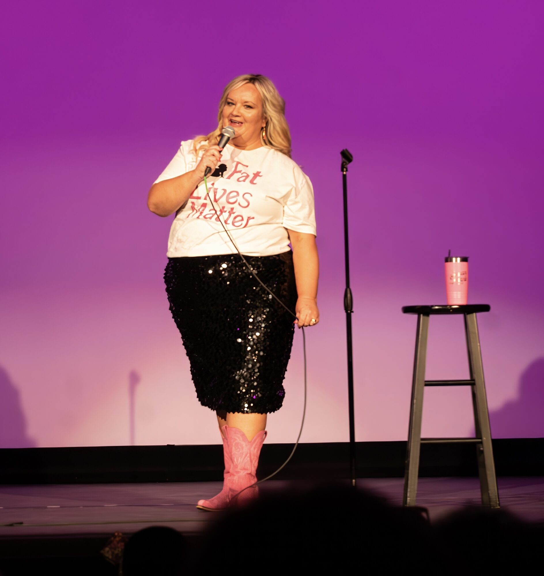 Woman on stage holding microphone, wearing a white T-shirt and black sequined skirt, pink boots, with a stool and water bottle nearby.