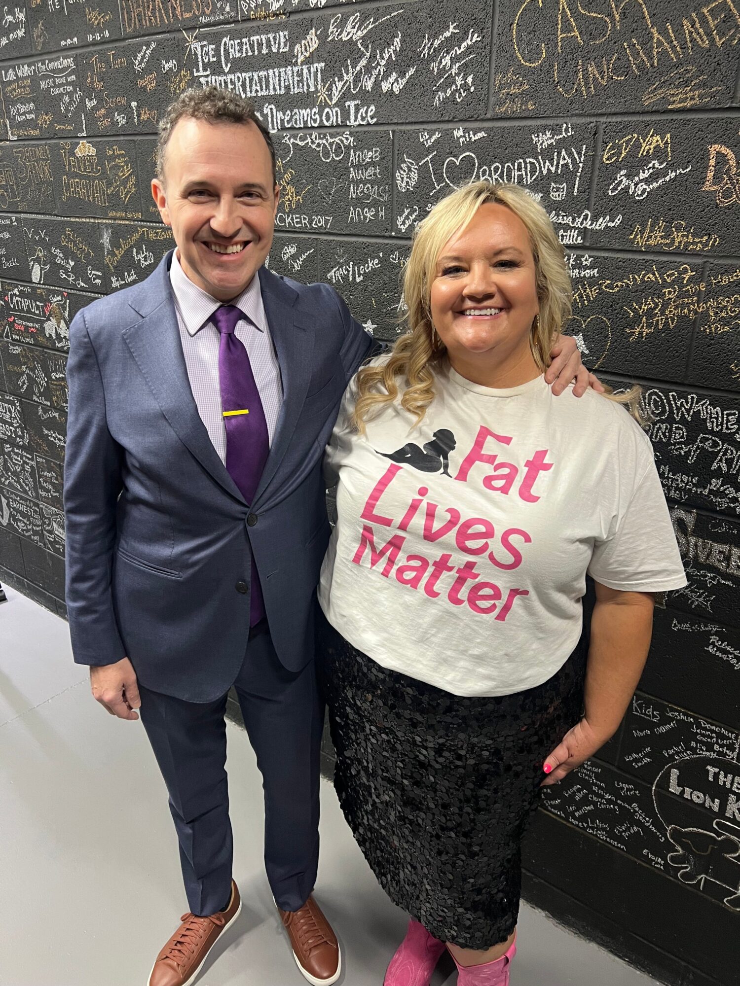 Two women standing together, one in a suit and the other in a white shirt with pink text, smiling.