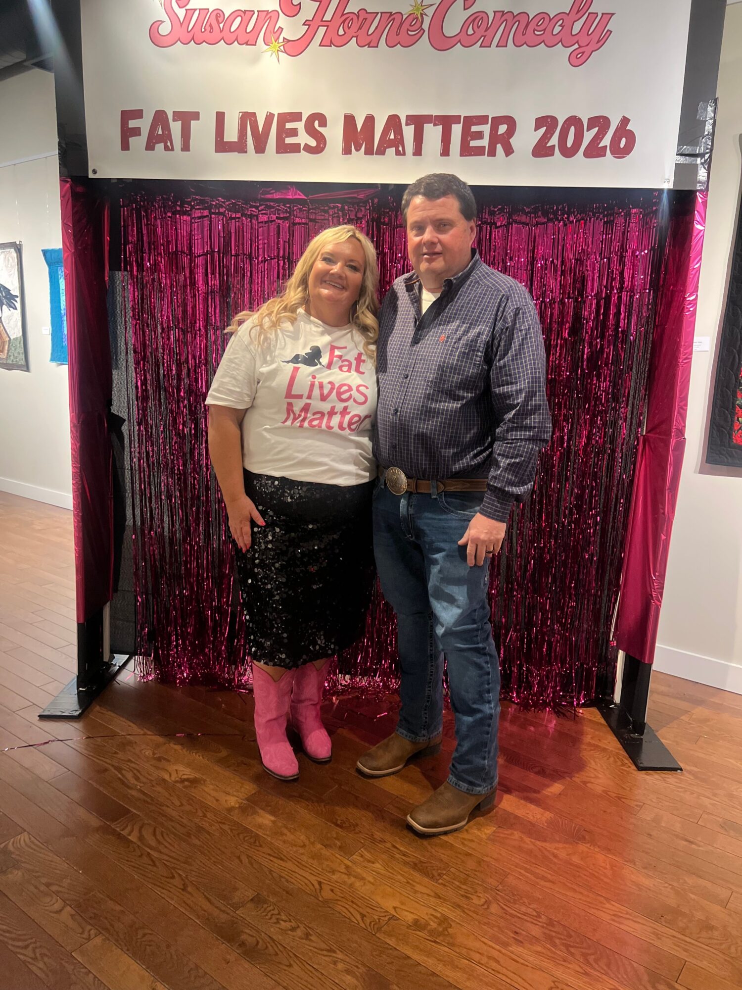 Two people standing in front of a pink curtain and a sign that reads 'FAT LIVES MATTER 2026'.