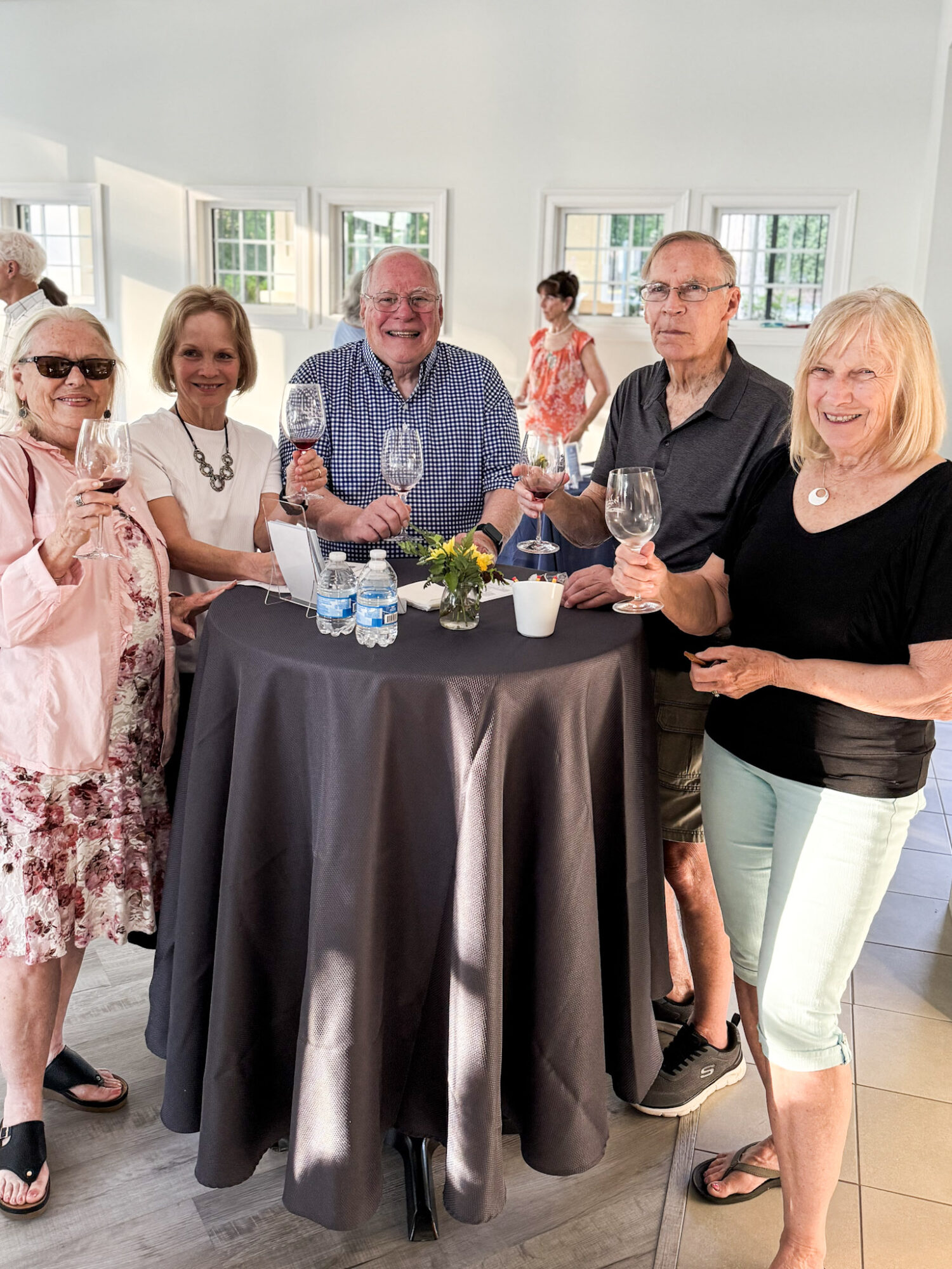 Group of six people standing around a high table with drinks, smiling at a social gathering indoors.
