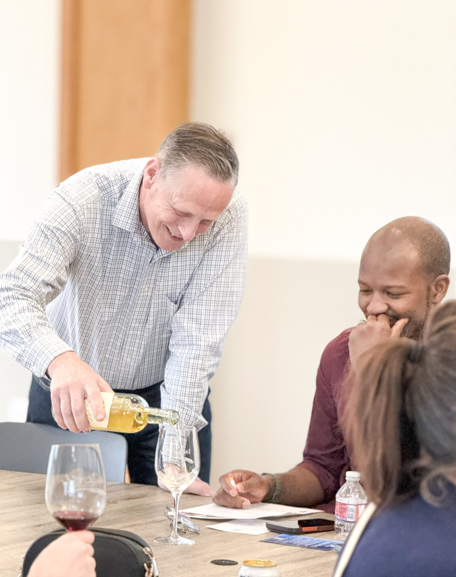 Man pouring wine at a table with people, one person smiling and covering mouth, others partially visible, in a bright room.