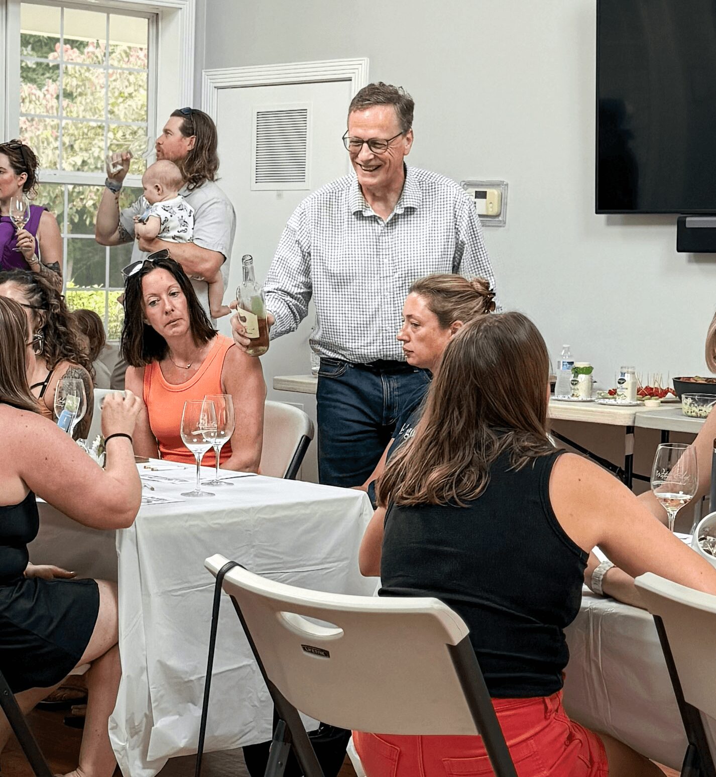 Group of people at a social gathering, some seated at a table, one standing and smiling, in a bright room.