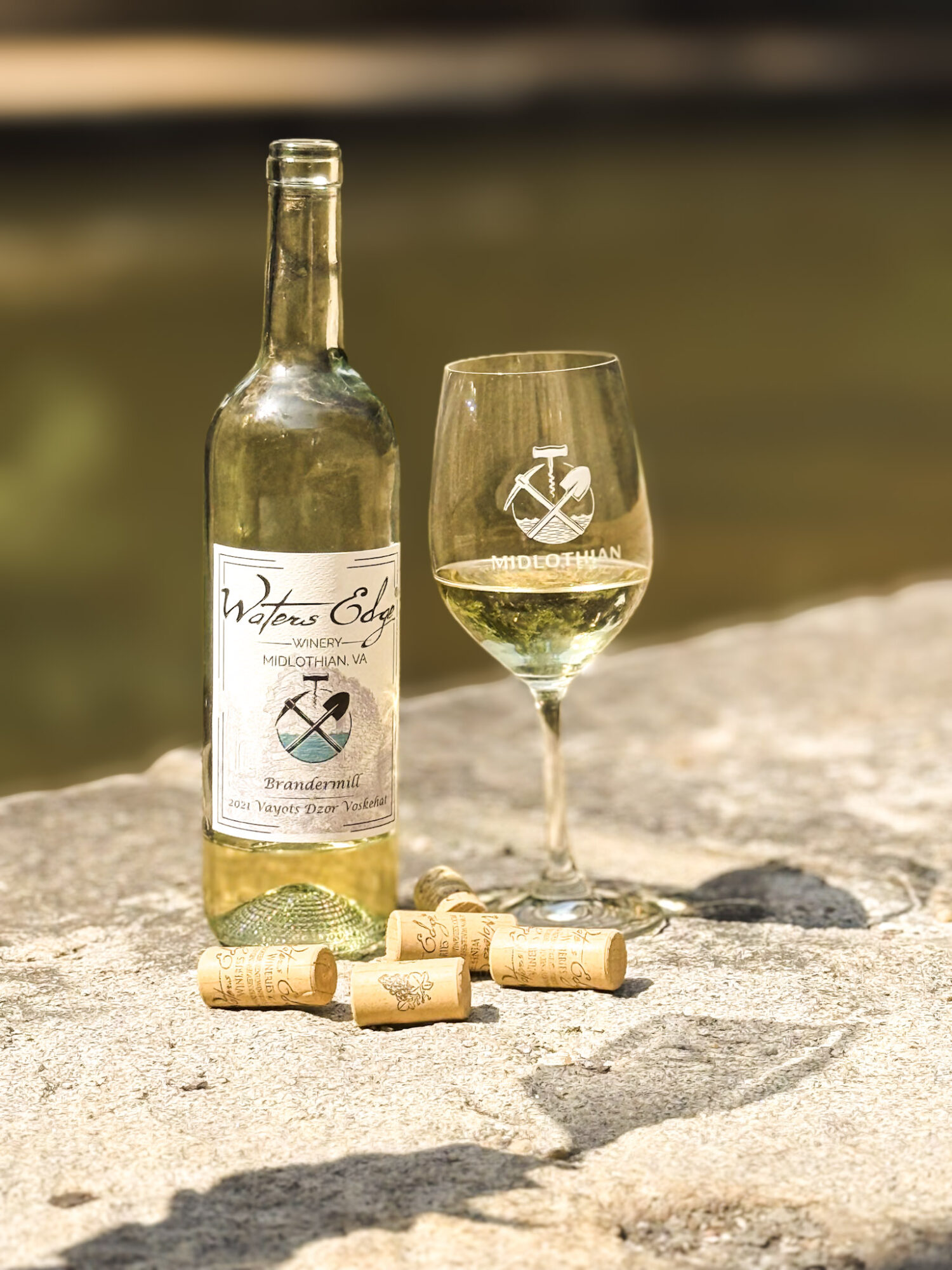 A wine glass and a bottle of white wine on a rock, with corks scattered around, outdoors near water.