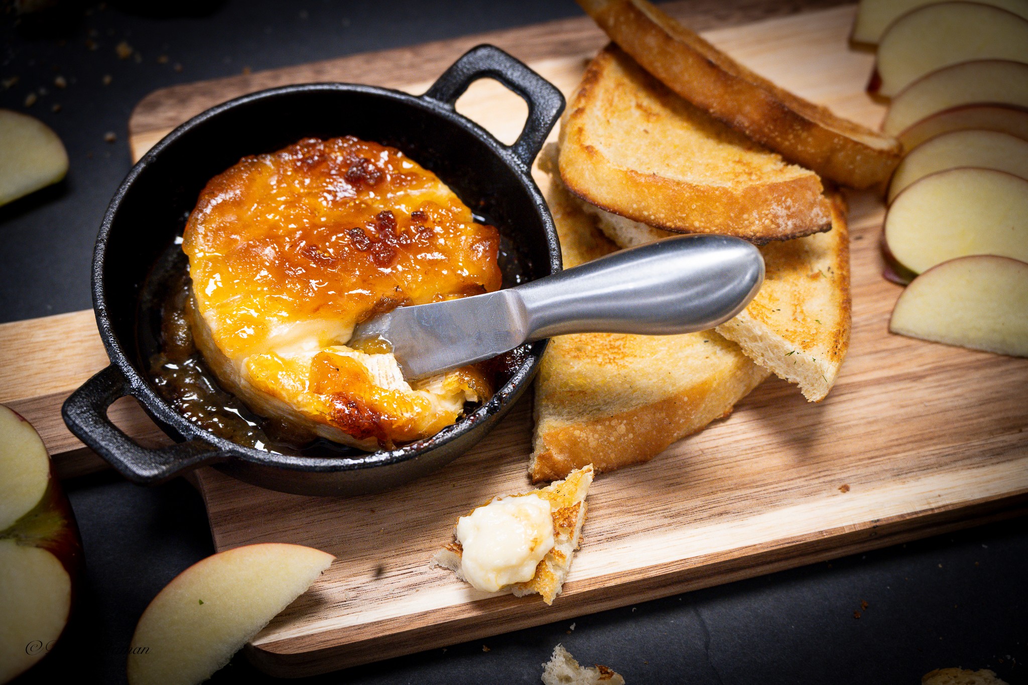Small black skillet with caramelized cheese, toasted bread slices, and sliced apples on a wooden board.