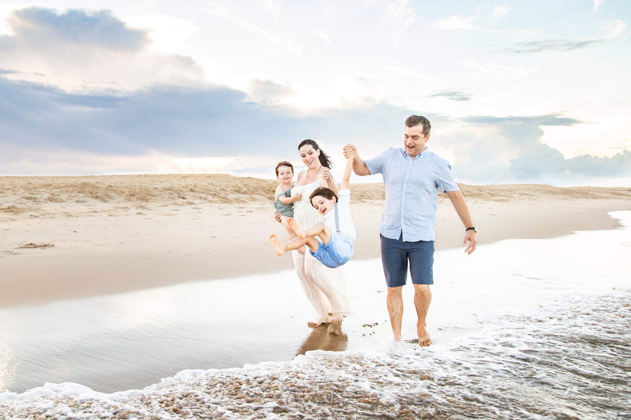 Family walking along the beach with children, holding hands, near the water, under a cloudy sky.