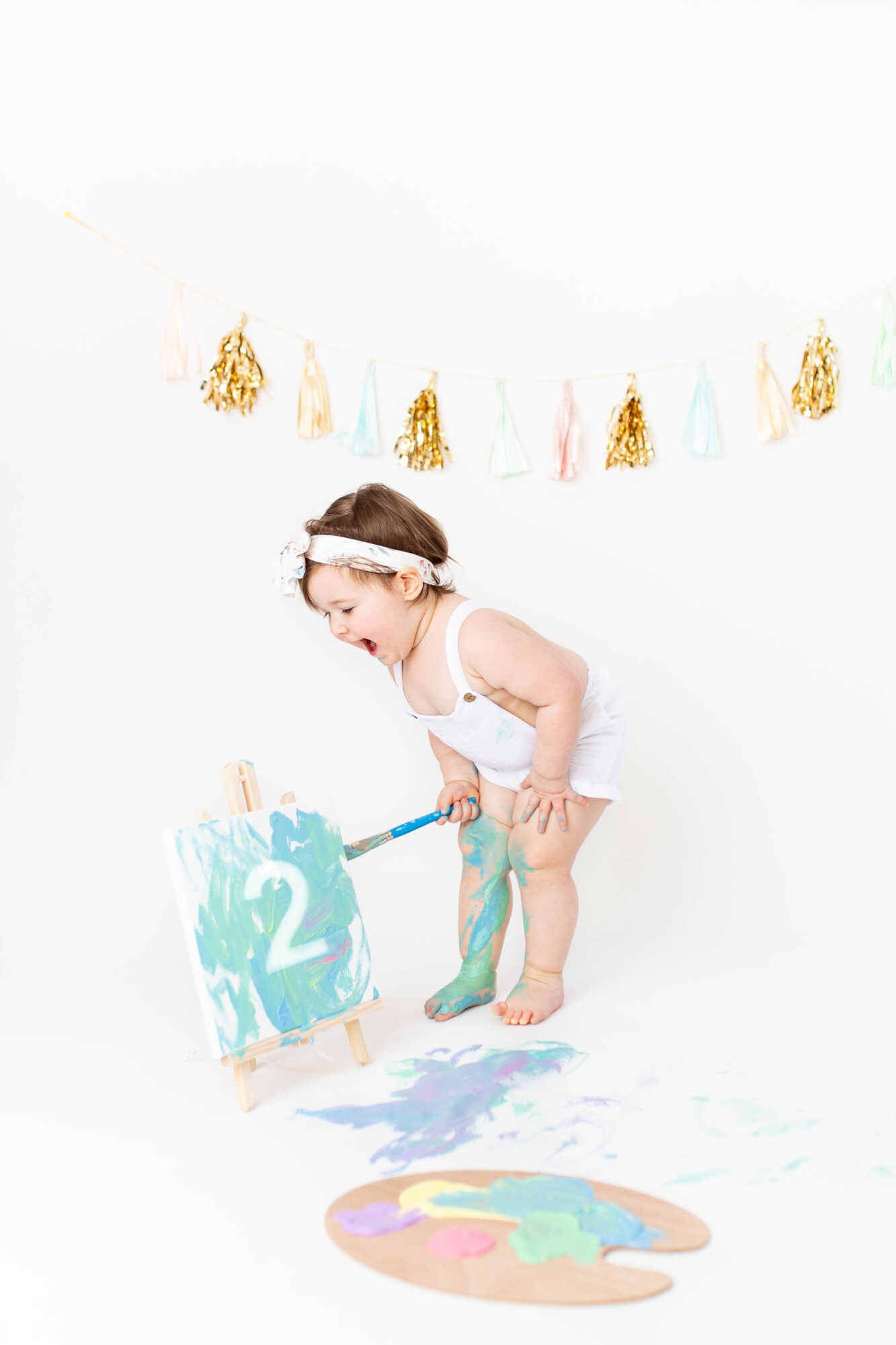 Child in white outfit with headband, playing with a paint palette and a small chalkboard with the number 2, colorful decorations above, white background.