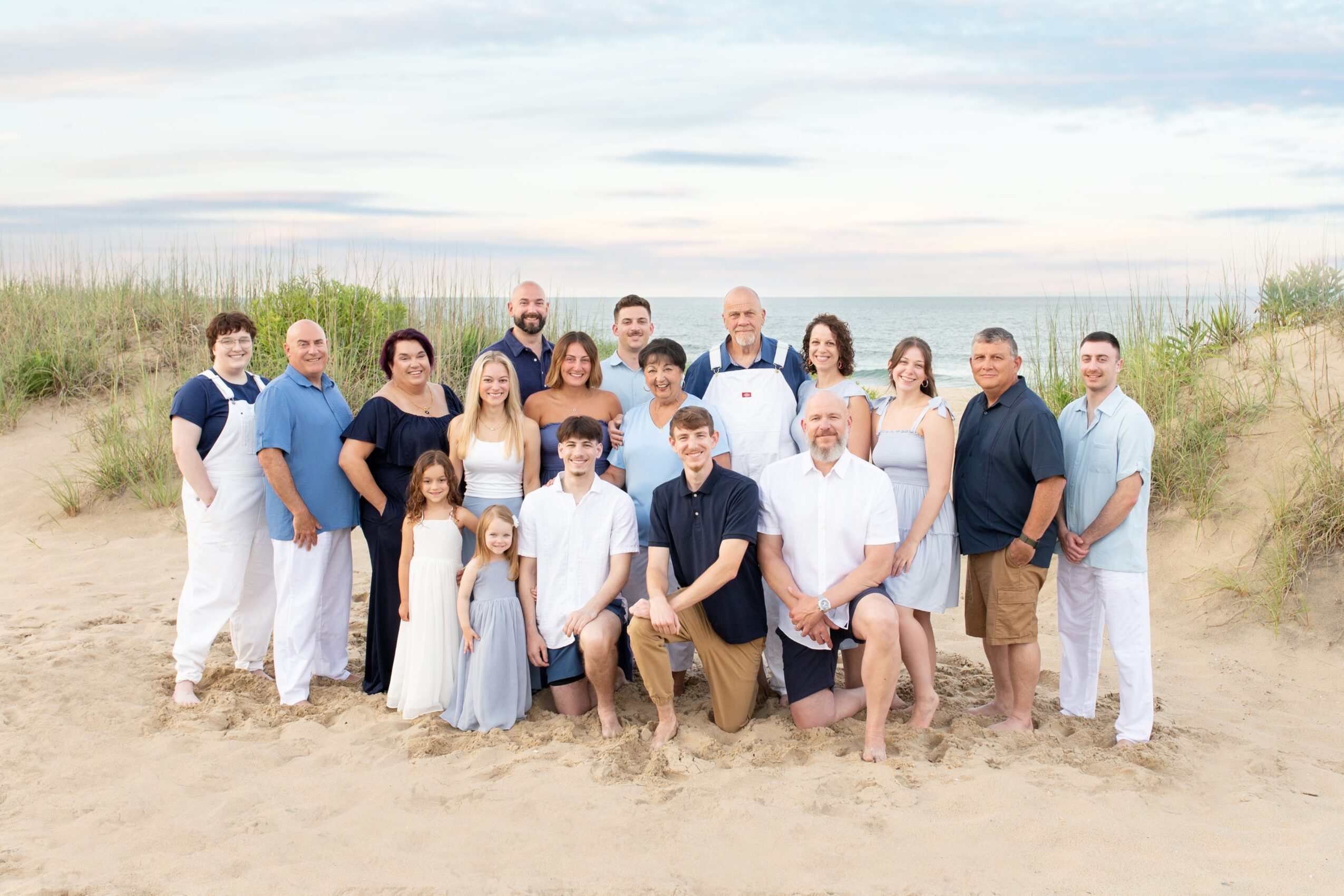 Group of 16 people standing on a sandy beach with grass and sky in background, smiling and posing for photo.