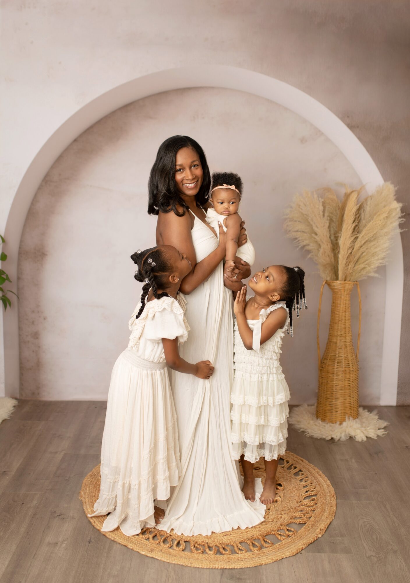 A woman with three children stands on a round rug in front of a white arch, smiling, with pampas grass in a vase nearby.