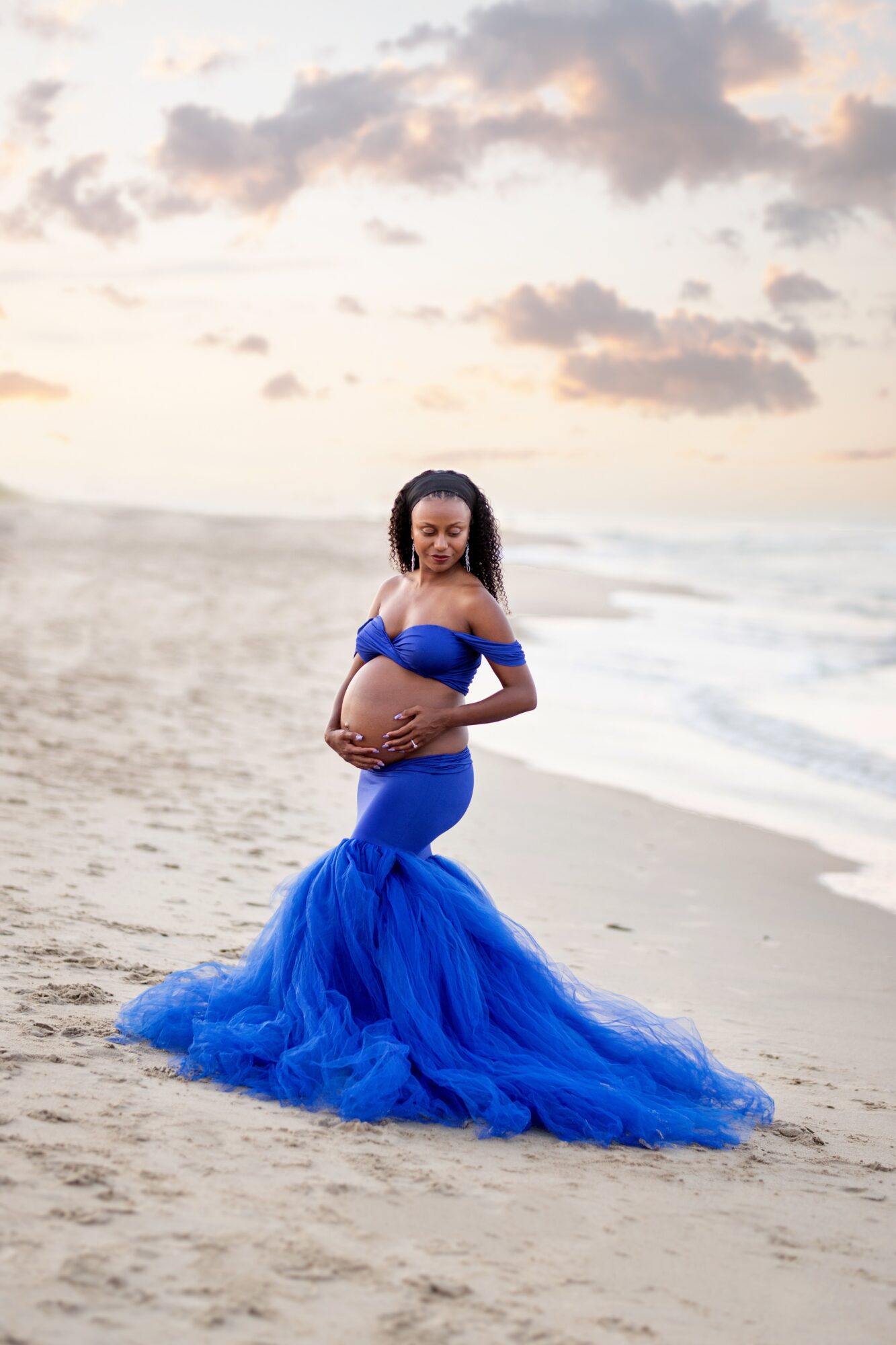 Pregnant woman in a blue dress standing on a beach with ocean waves and cloudy sky in the background.