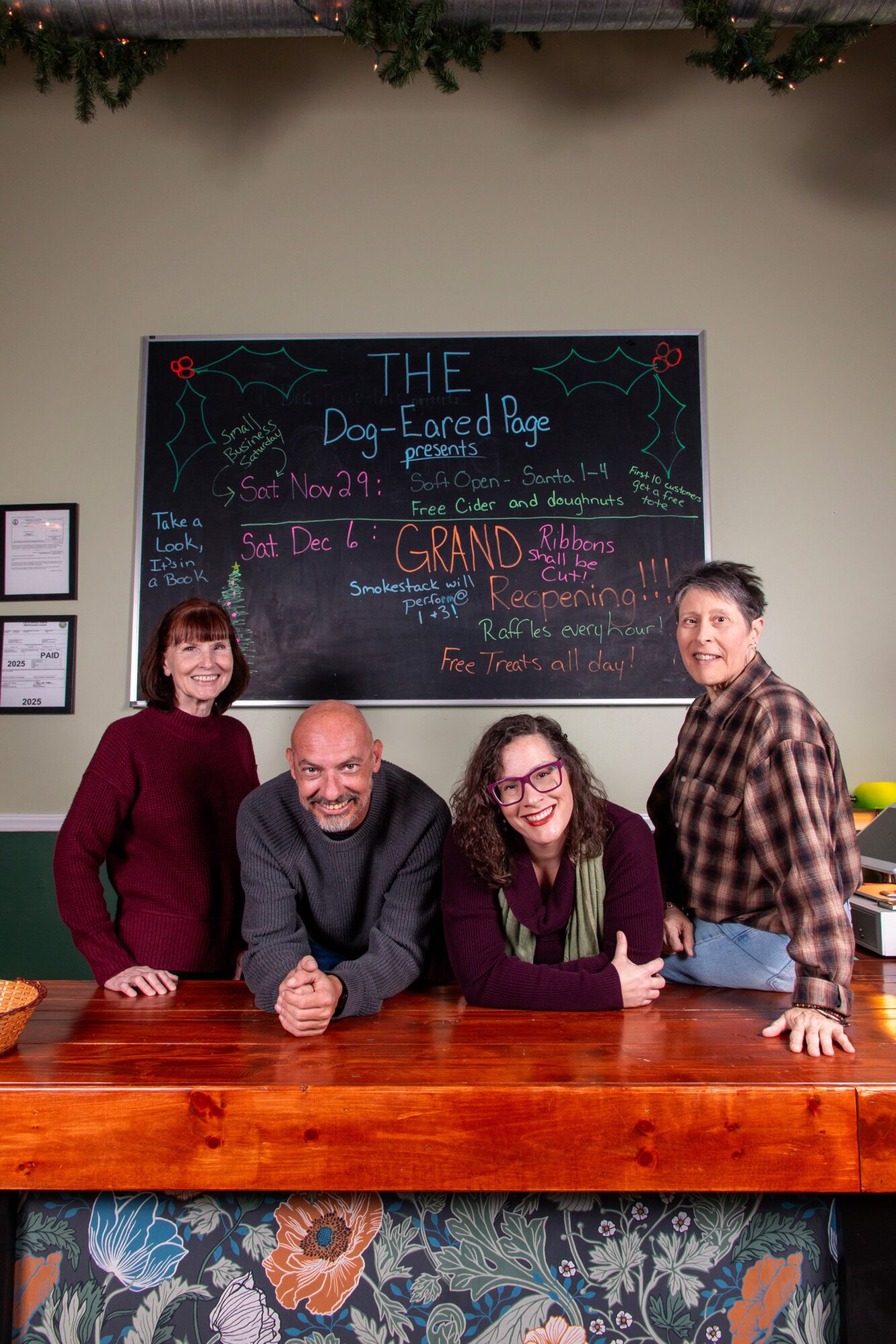 Four people stand behind a wooden counter in front of a chalkboard with colorful writing, smiling at the camera.