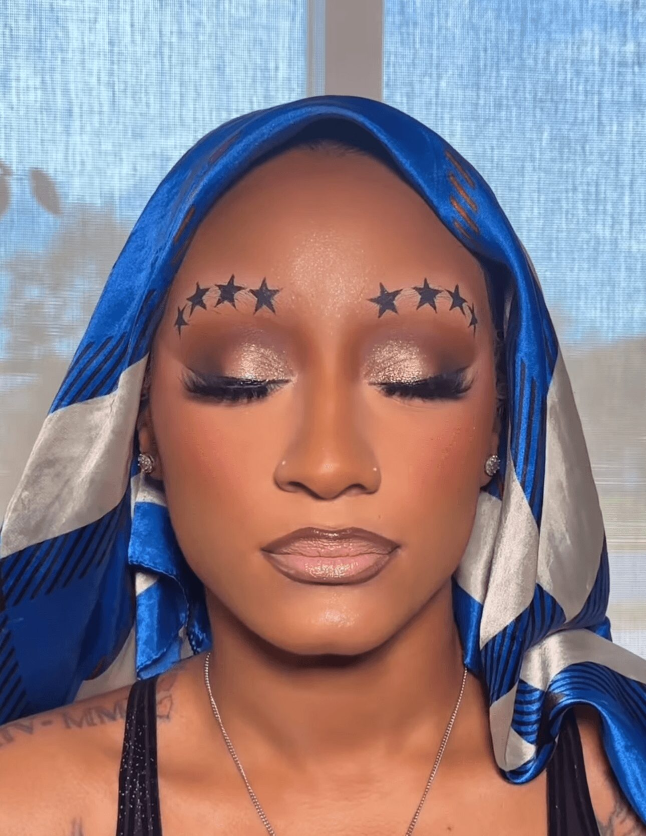 Woman with closed eyes, wearing a blue and white headscarf, makeup, and star-shaped face decorations.