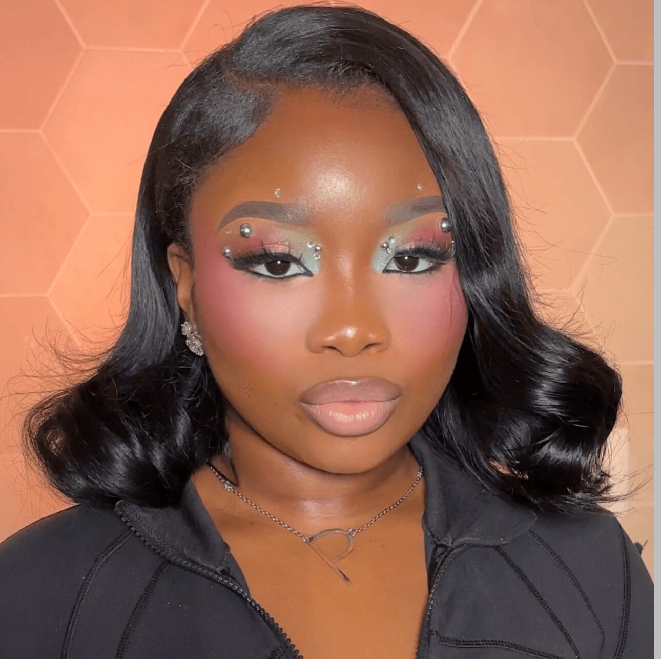 Woman with styled black hair, colorful makeup, earrings, and a black top, standing against a hexagonal patterned background.