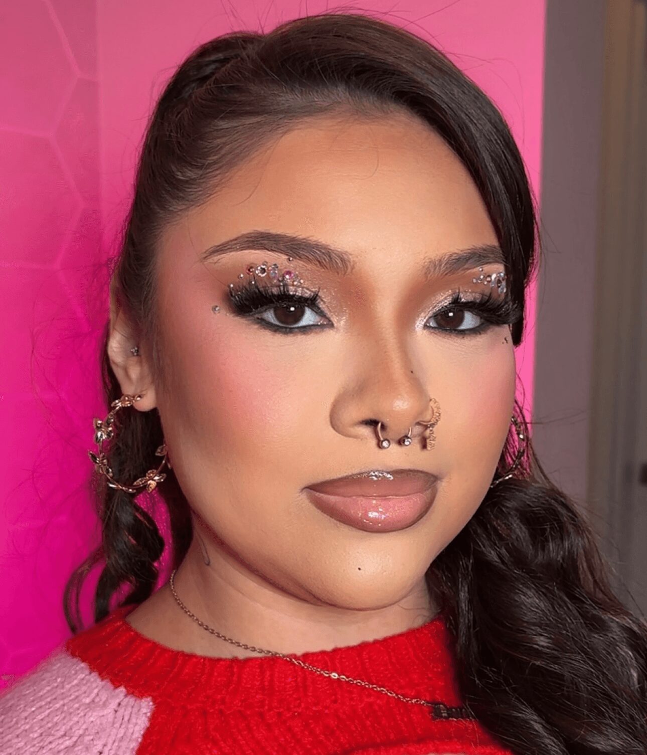 Close-up of a woman with makeup, glitter eyeshadow, septum piercing, and long dark hair, wearing a red top and gold earrings.