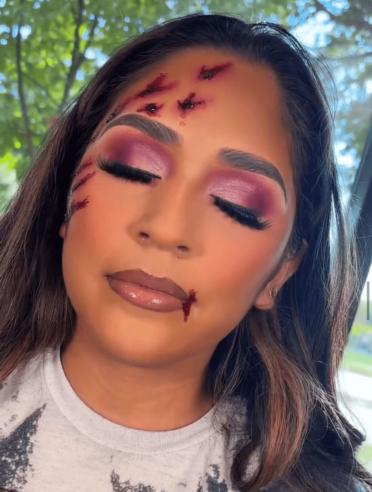 Woman with makeup and fake scars on face, eyes closed, outdoors with trees in background.