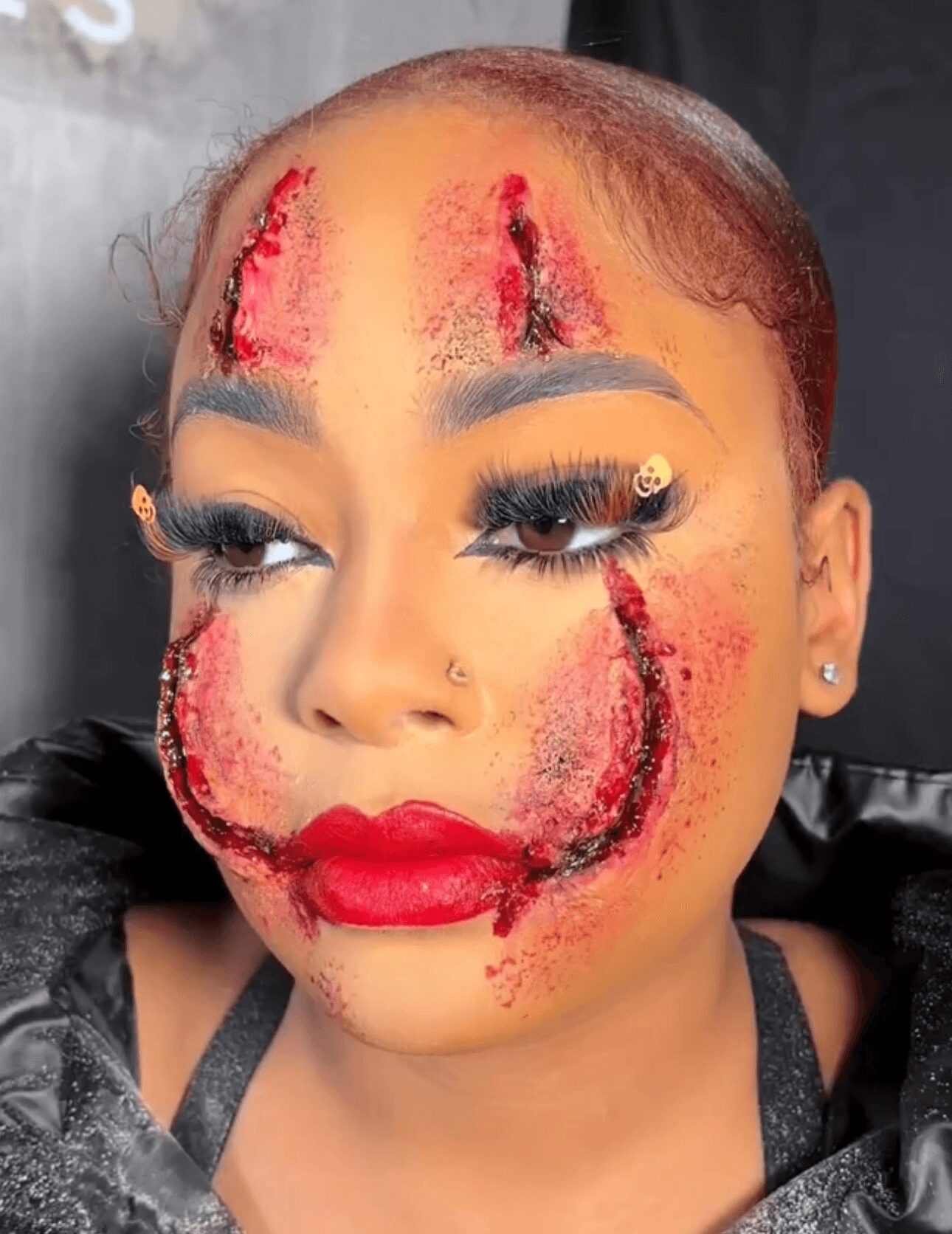 Close-up of a woman with makeup and special effects, including red and black streaks on her face, red lipstick, and long eyelashes.