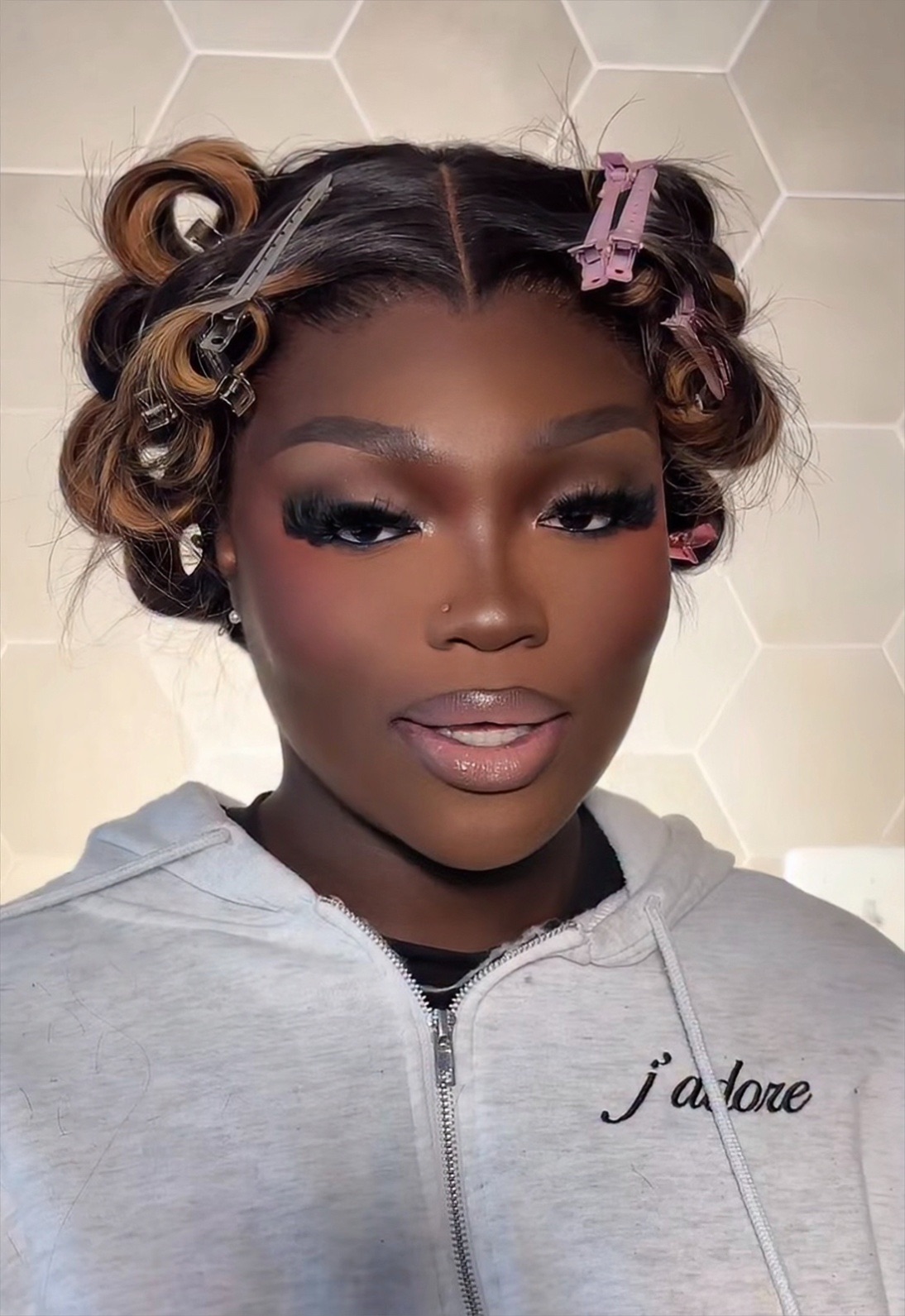Close-up of a woman with styled hair and makeup, wearing a light gray hoodie with text, standing against a hexagonal wall.