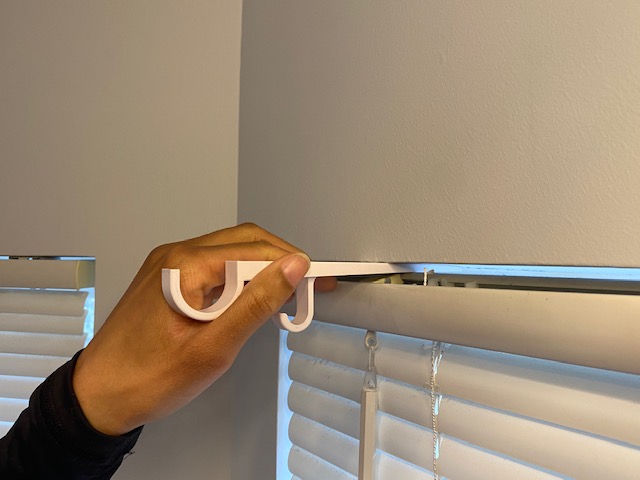 Hand adjusting horizontal window blinds with a white cord, wall and window visible in background.