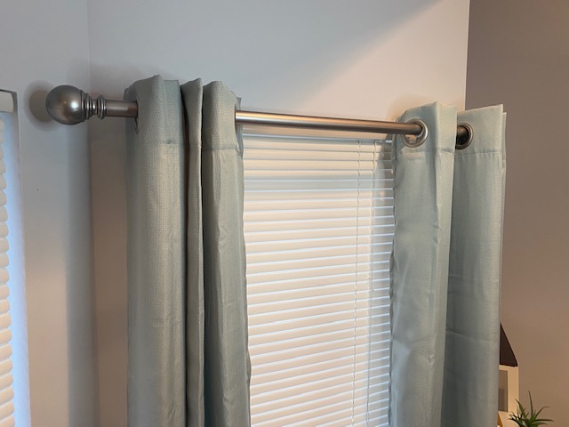 Curtains hanging on a metal rod in front of a window with blinds, wall on the left and a small plant on the right