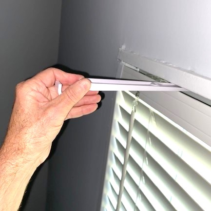 Hand holding a tablet near window blinds, viewed from the side, with a wall in the background.