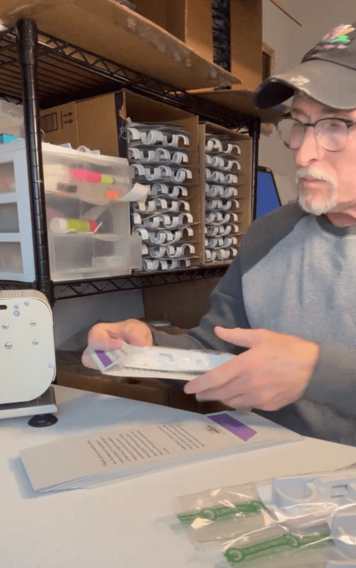 Man wearing glasses and a cap working at a desk with organized supplies and a computer monitor.