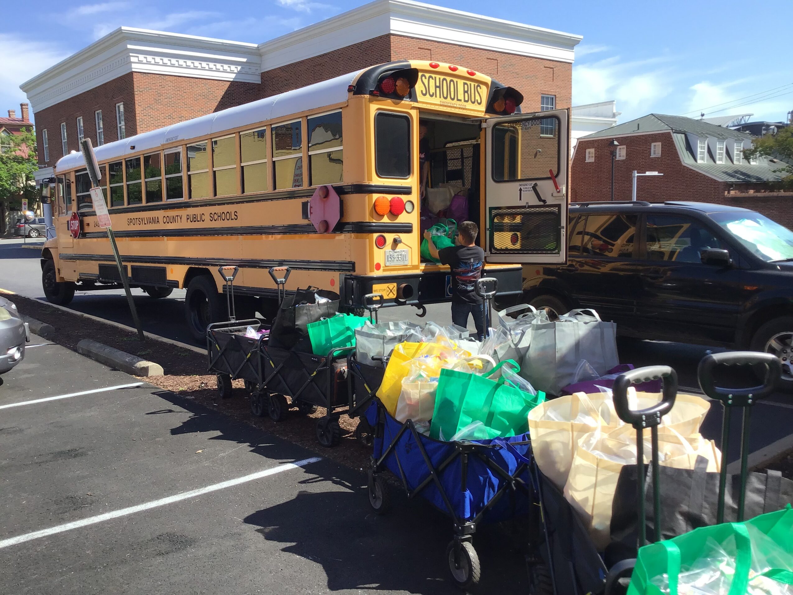 School bus parked in lot with luggage carts and children nearby, brick building in background, clear sky.