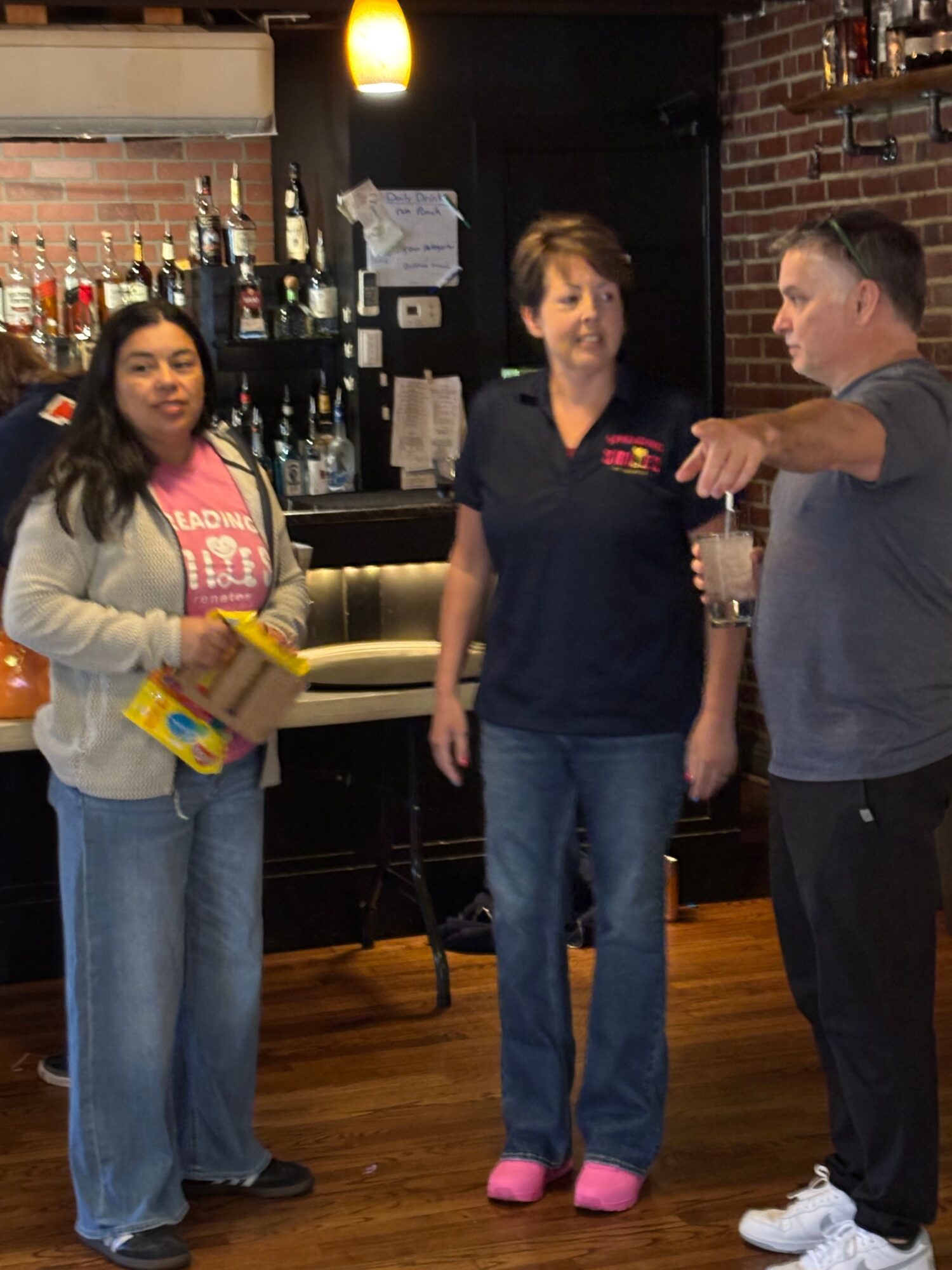 Three people standing in a bar, two women and one man, with bar shelves and bottles in the background.