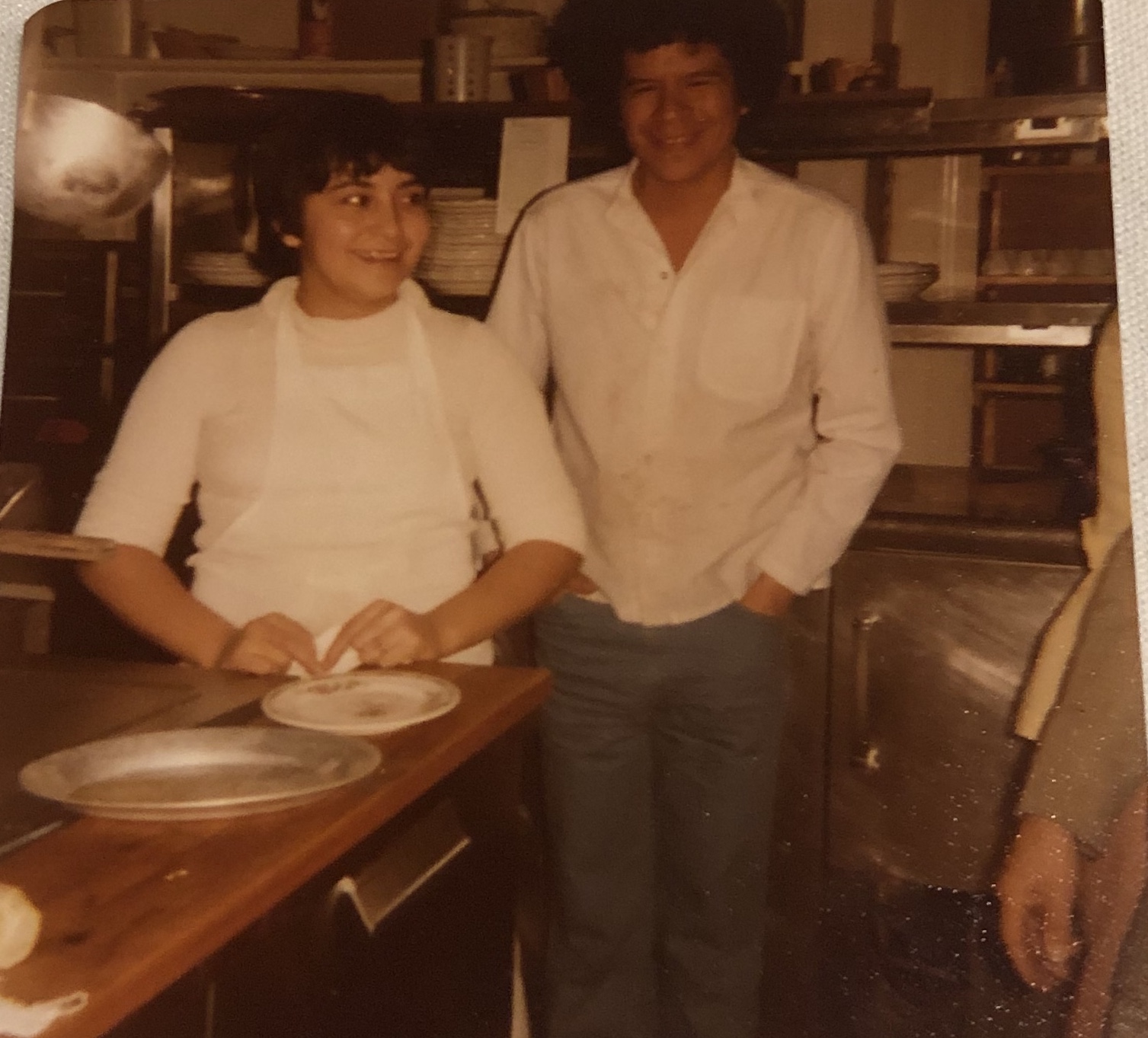Two people standing in a kitchen, smiling, with plates on a wooden counter, one wearing a white apron and the other in a white shirt.