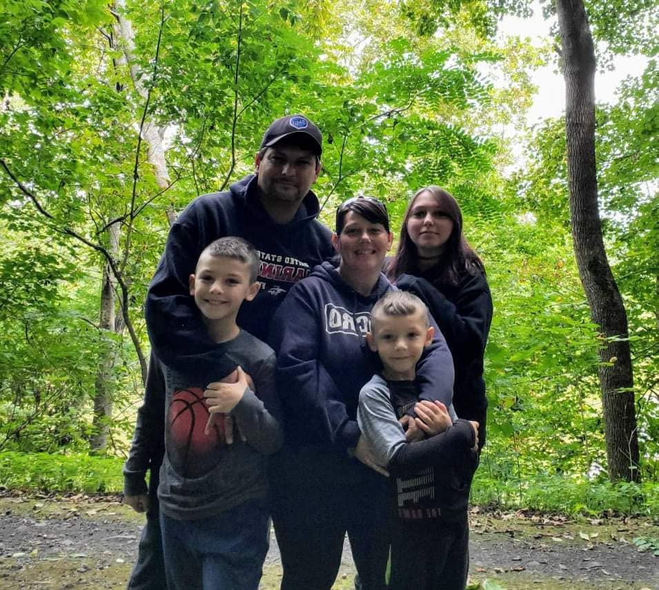 Group of five people, two adults and three children, standing outdoors in a green forest, smiling at the camera.