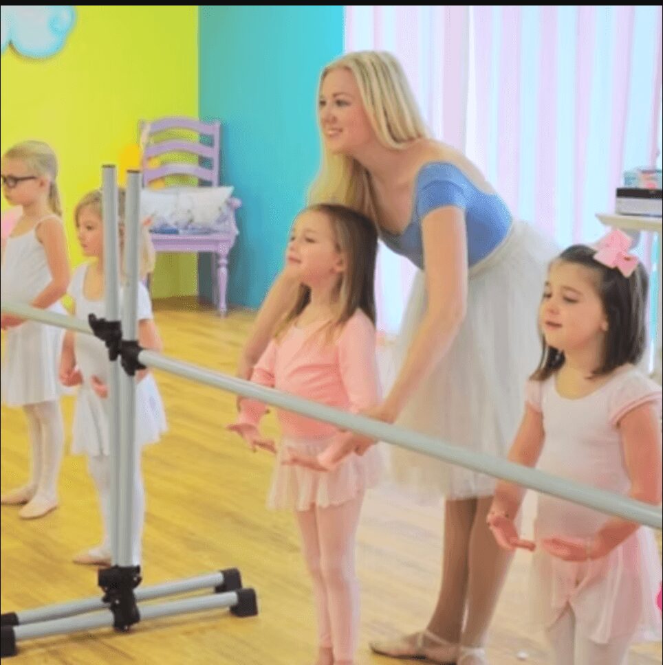 A woman and children in a colorful room, with a woman guiding a child holding a sword, children in ballet costumes.