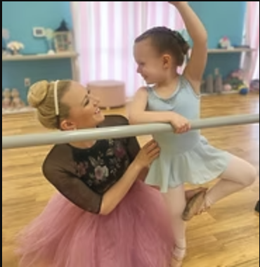 A ballet instructor and young girl practicing at a barre in a dance studio, smiling and engaging with each other.
