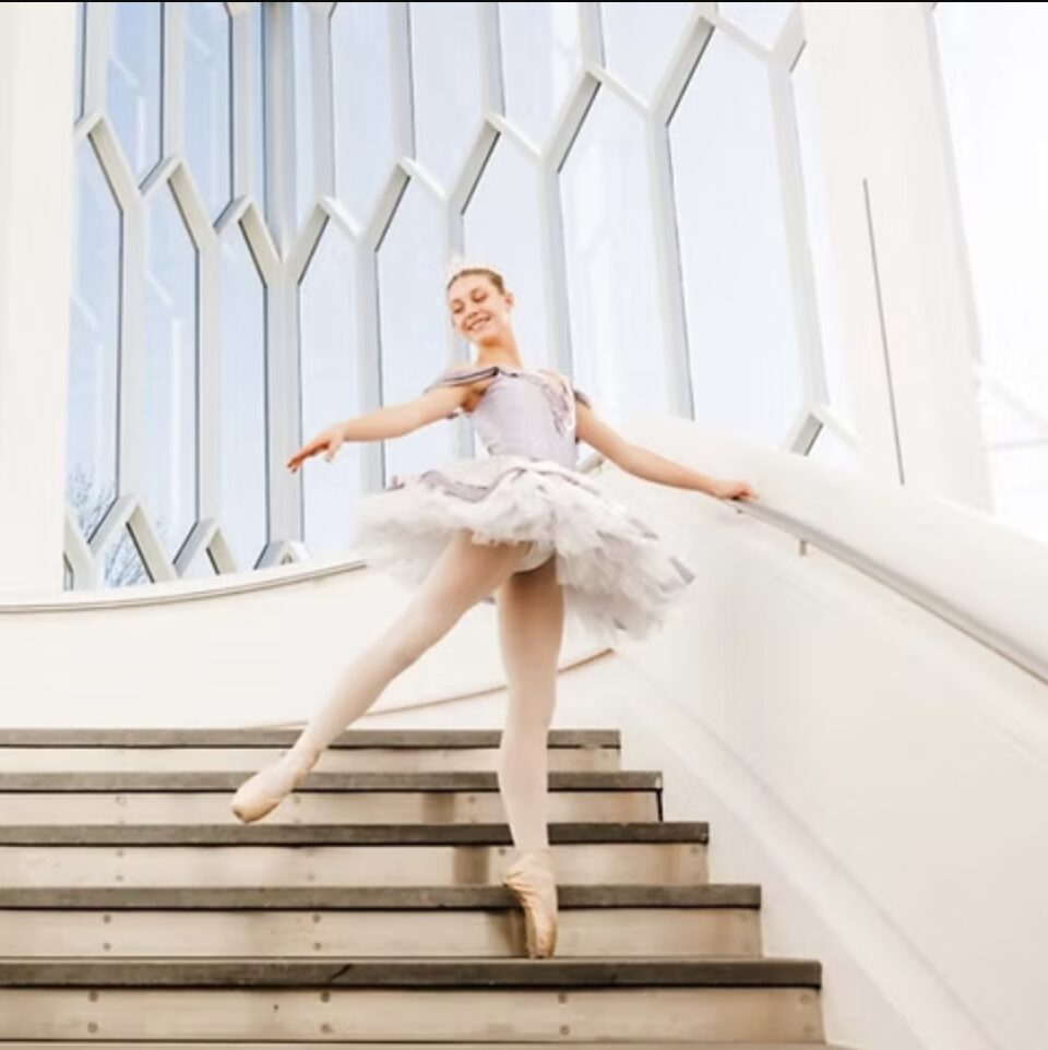 Ballet dancer in tutu on stairs, arms extended, smiling, with large windows behind her.