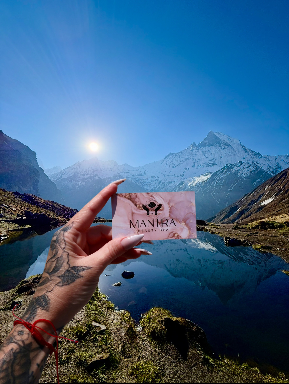 Person holding a pink card with a mountain landscape and lake in the background, clear blue sky, snow-capped peaks.