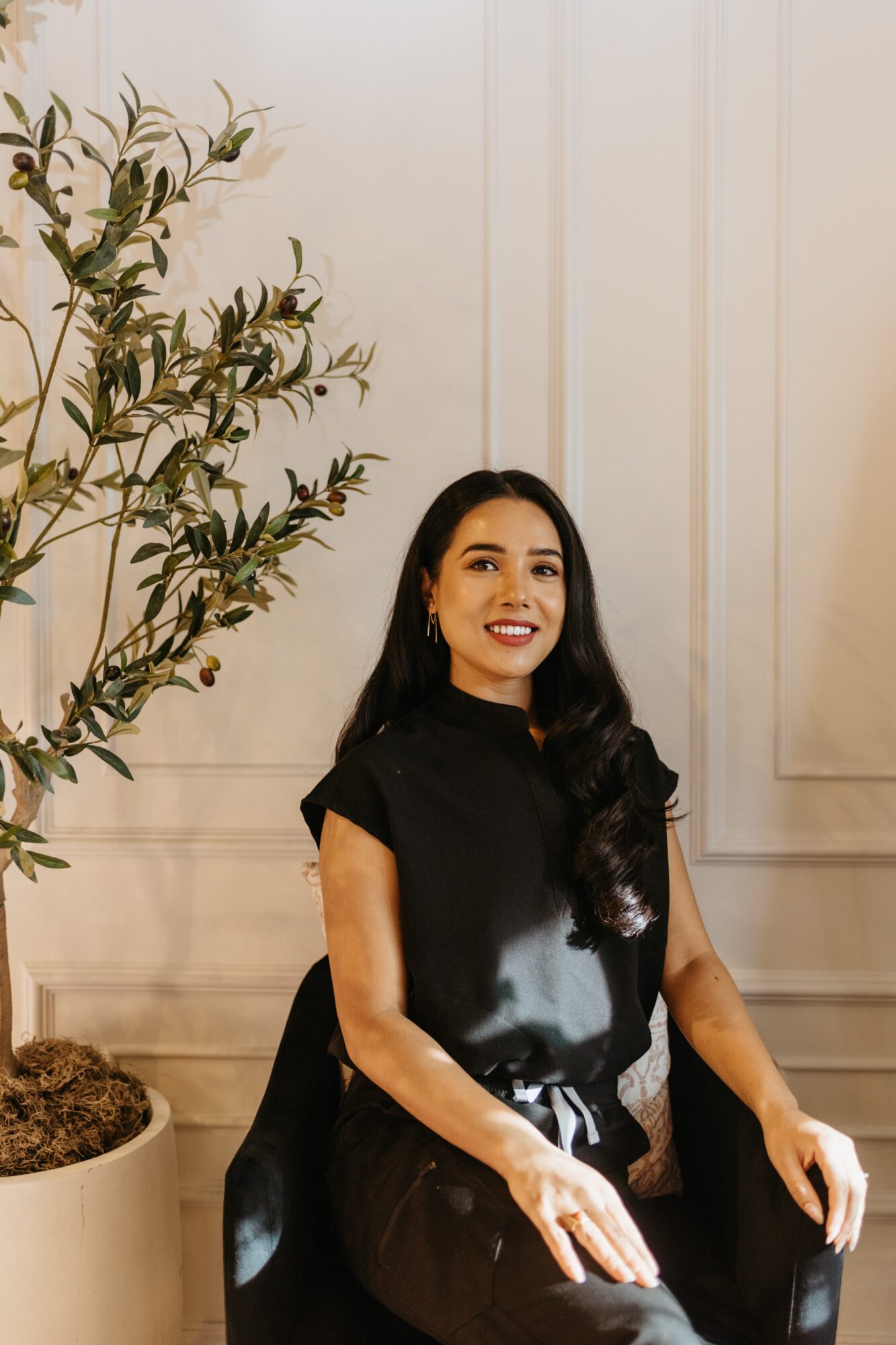 Woman with long dark hair wearing black outfit, smiling, sitting indoors near a potted plant, cream-colored wall background.