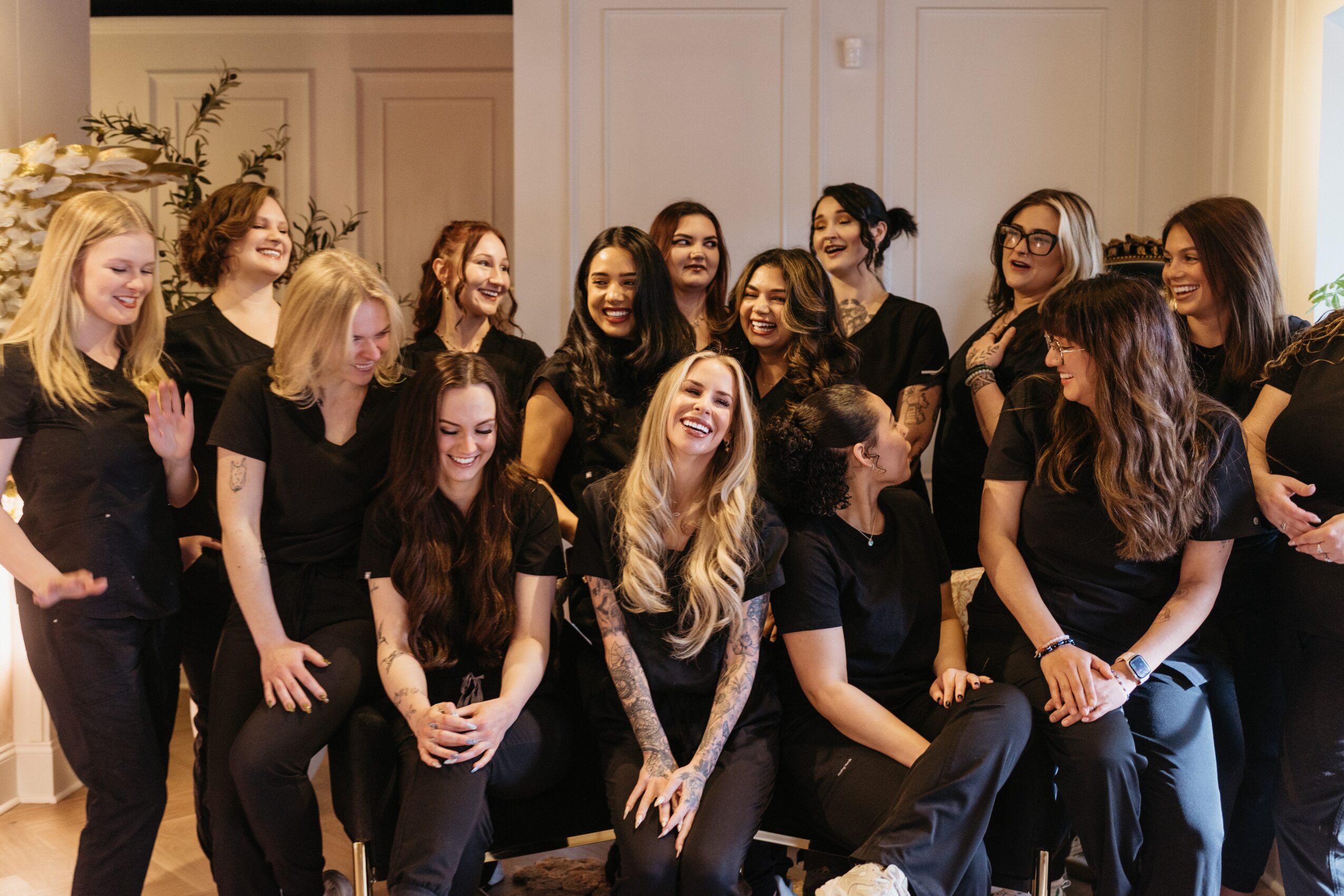Group of women in black shirts smiling and laughing indoors, some standing and some sitting, in a well-lit room.