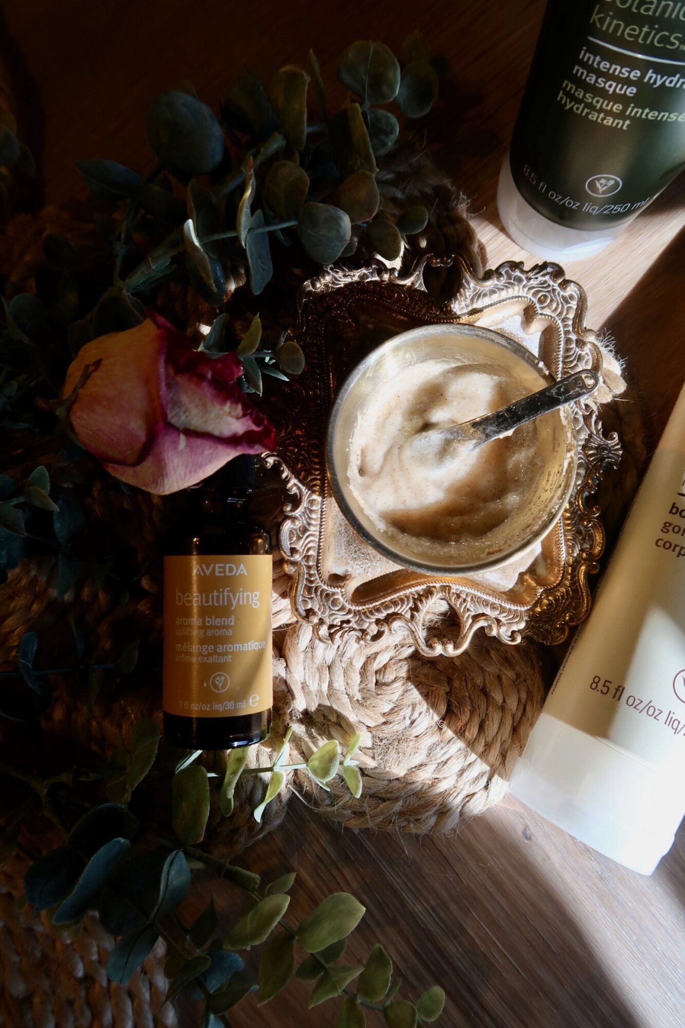 A glass cup with a spoon inside, placed on a decorative silver tray, surrounded by skincare products and eucalyptus leaves.
