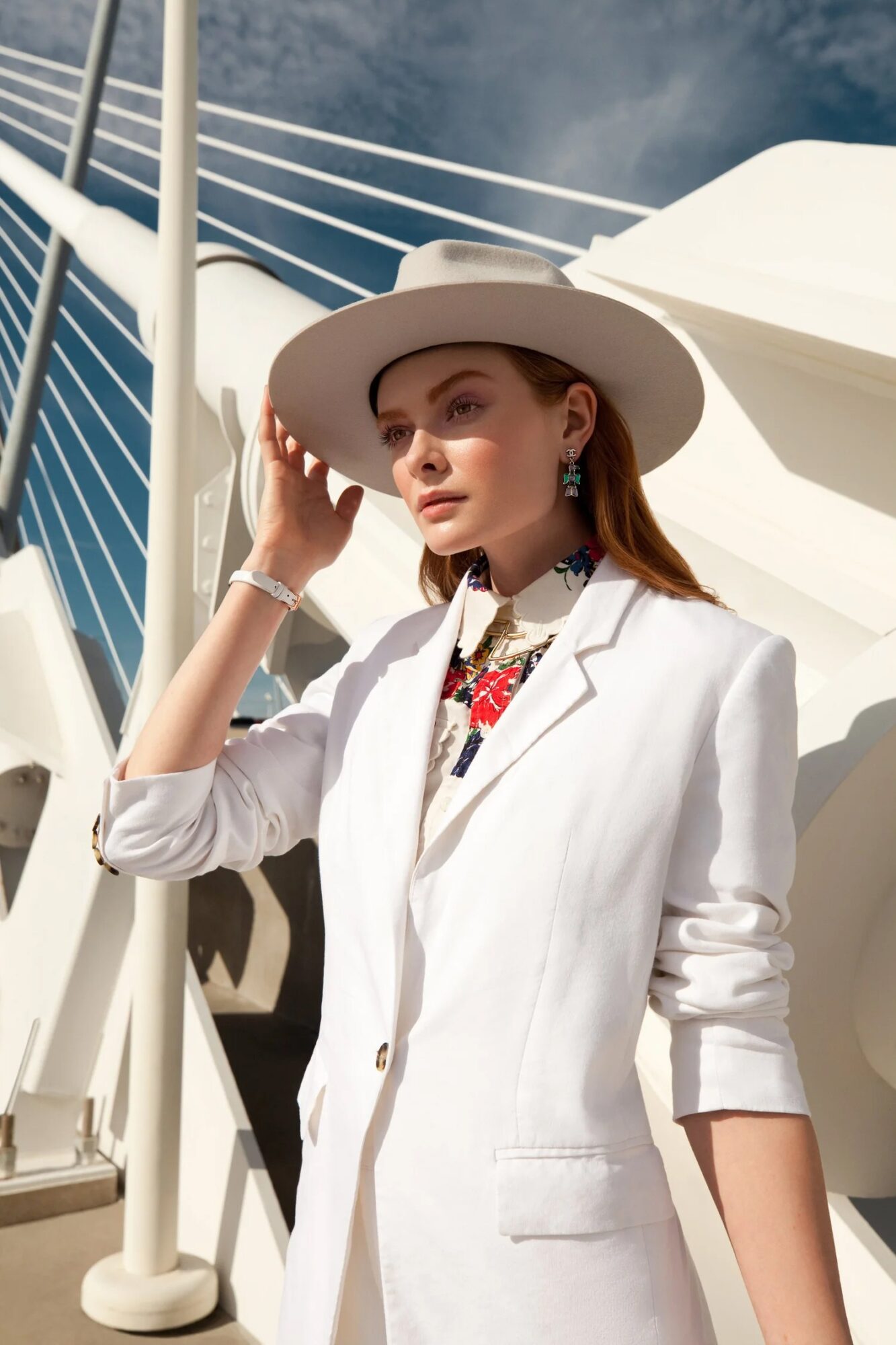 Woman in white blazer and wide-brimmed hat standing outdoors near a modern architectural structure.