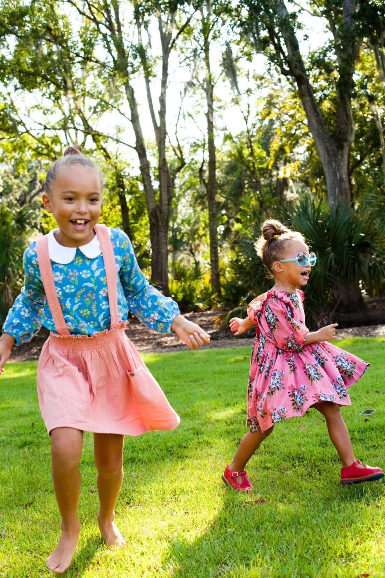 Two young girls playing outdoors on grass, holding hands, smiling, with trees in the background.